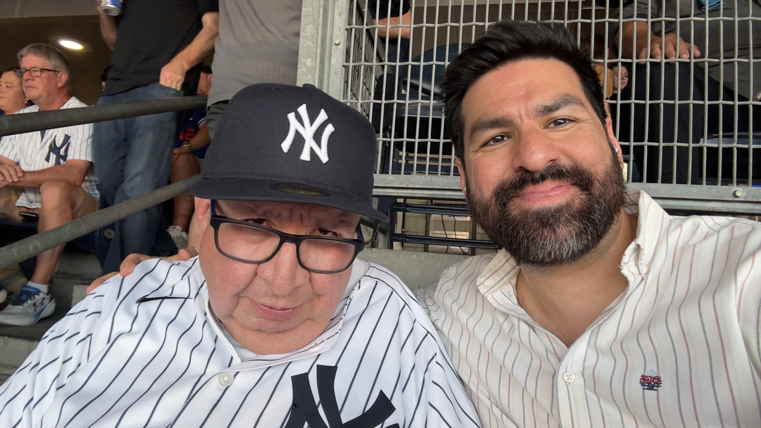 Two men at a baseball game taking a selfie. One man is wearing a New York Yankees cap and glasses, the other has dark hair and a beard. They are sitting in stadium seats with other spectators in the background.