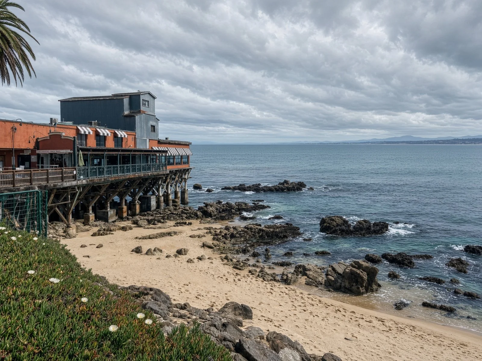 A seaside scene with a wooden pier or deck extending over rocks and sand, positioned near a beach, with a building on stilts, and a cloudy sky overhead.