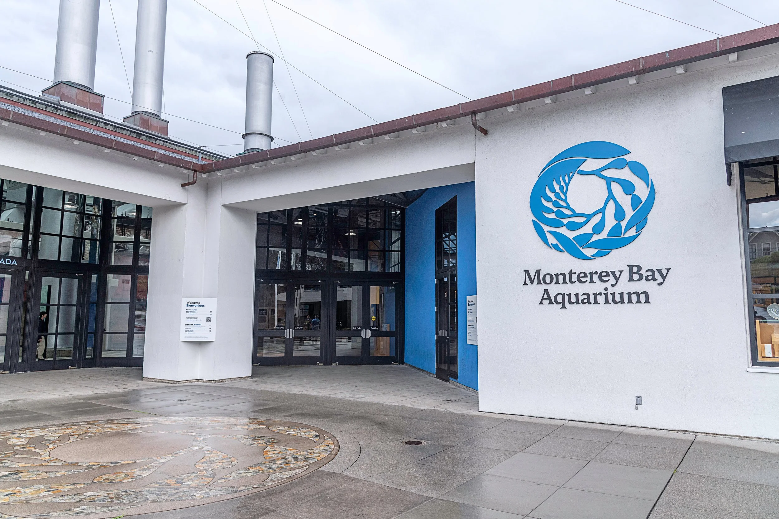 Entrance to Monterey Bay Aquarium with a blue logo on the white building wall and glass doors.