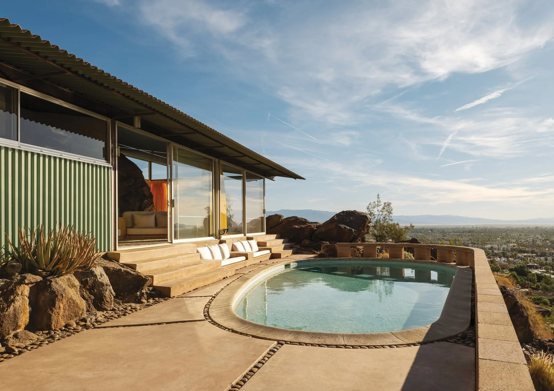 A residential house with large glass windows and a corrugated metal roof, situated on a hillside with a swimming pool in the foreground, desert landscape, and cityscape in the distance under a partly cloudy sky.