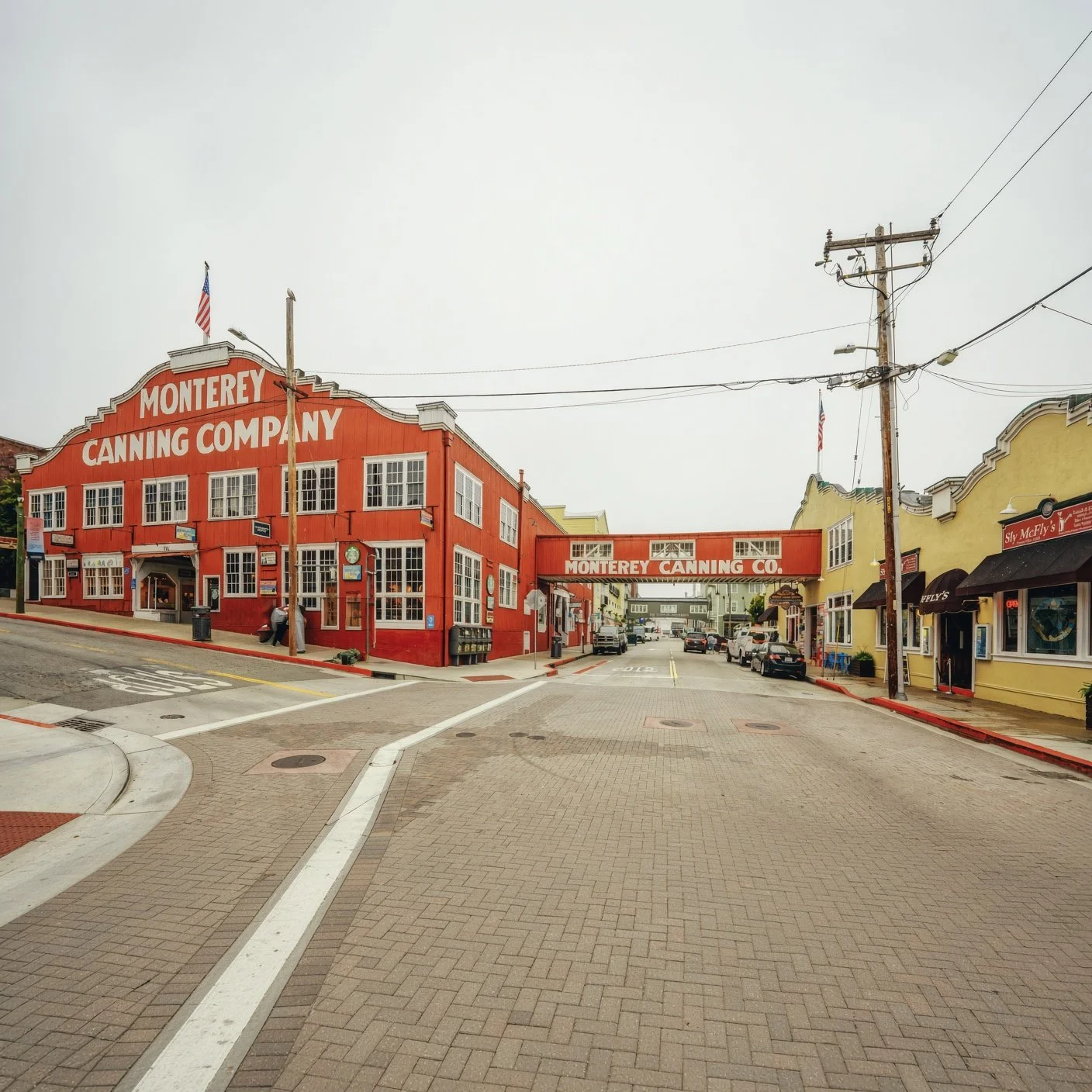 A street scene in Monterey, California, with a red building labeled 'Monterey Canning Company' and a yellow building to the right, both with storefronts and parked cars along the street.