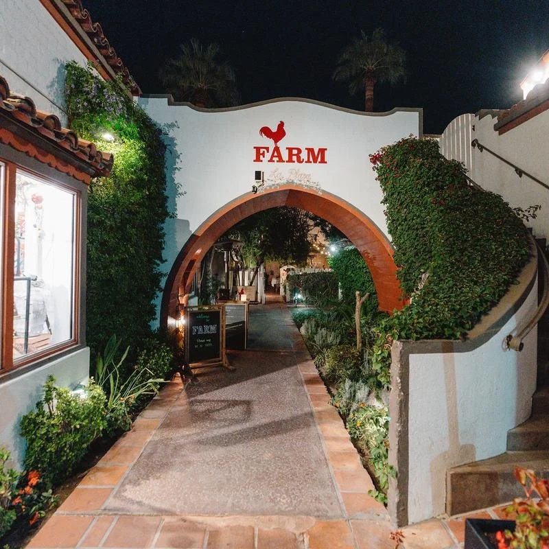 Night view of the entrance to a farm-themed establishment with an archway, decorative plants, and the word 'FARM' with a rooster on top of a white wall.