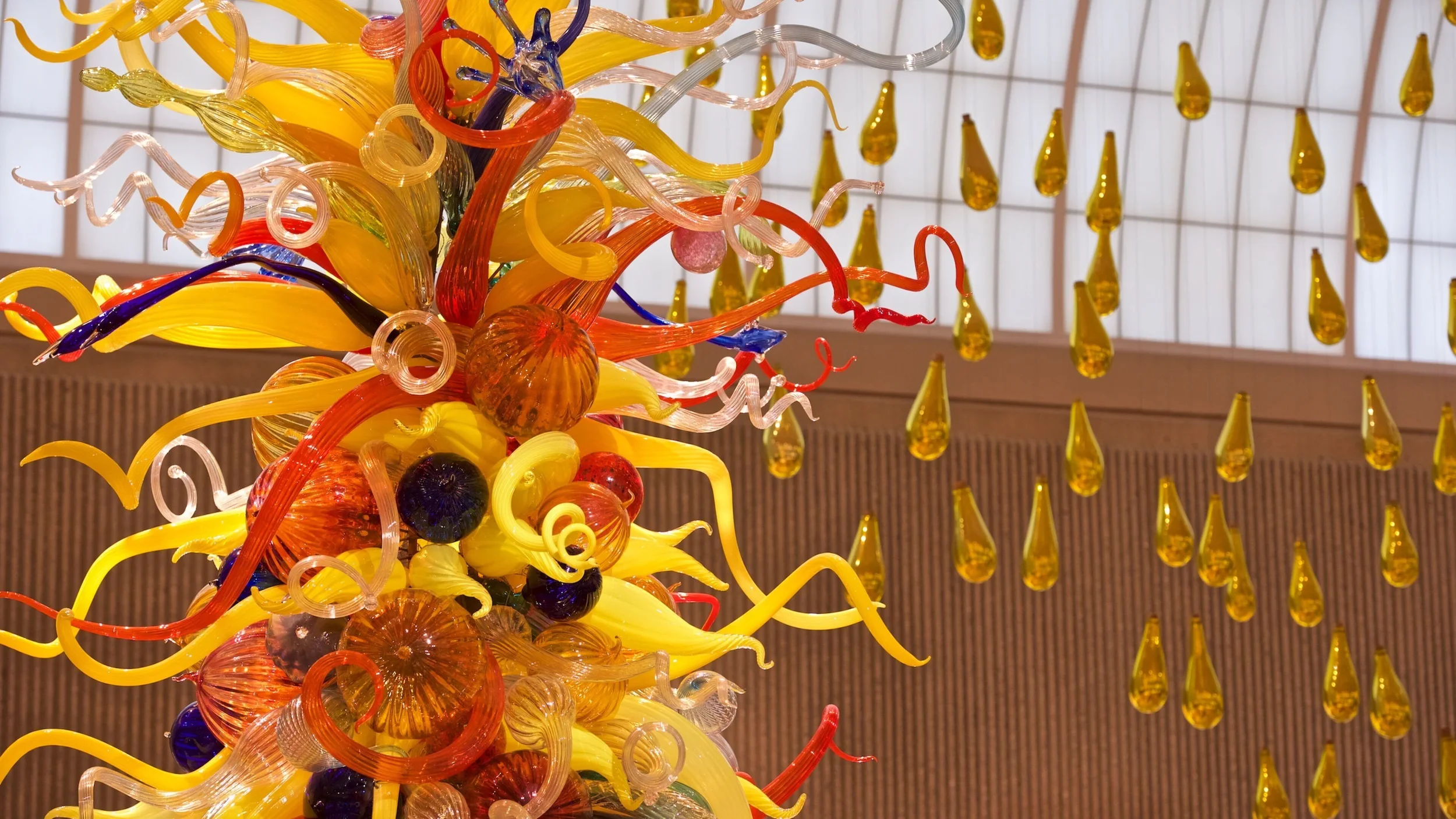 Colorful glass sculpture with various shaped swirls and orbs, hanging in front of a window with yellow glass drop ornaments.