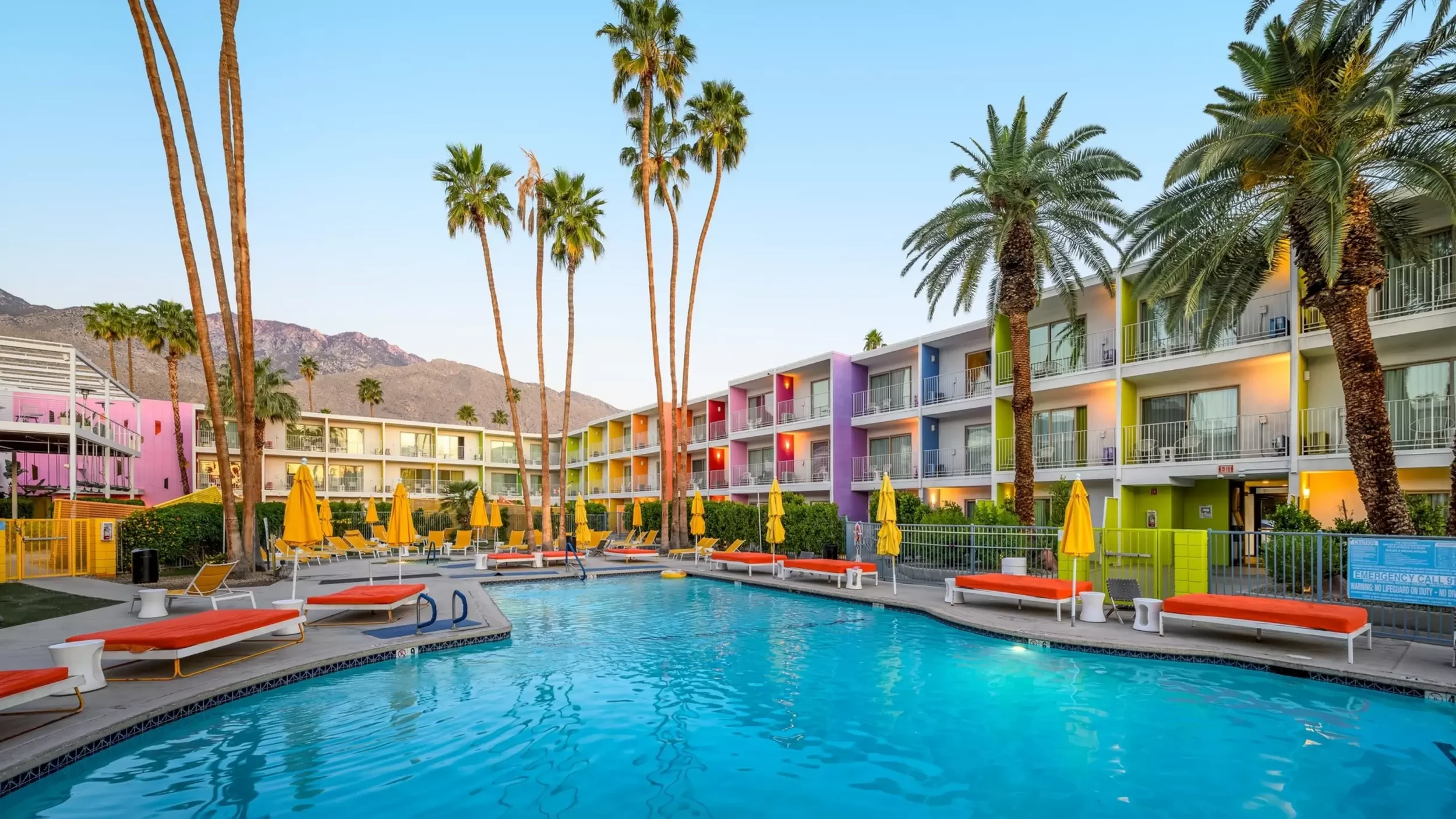 Colorful hotel with a swimming pool surrounded by orange lounge chairs and yellow umbrellas, tall palm trees, and mountains in the background.