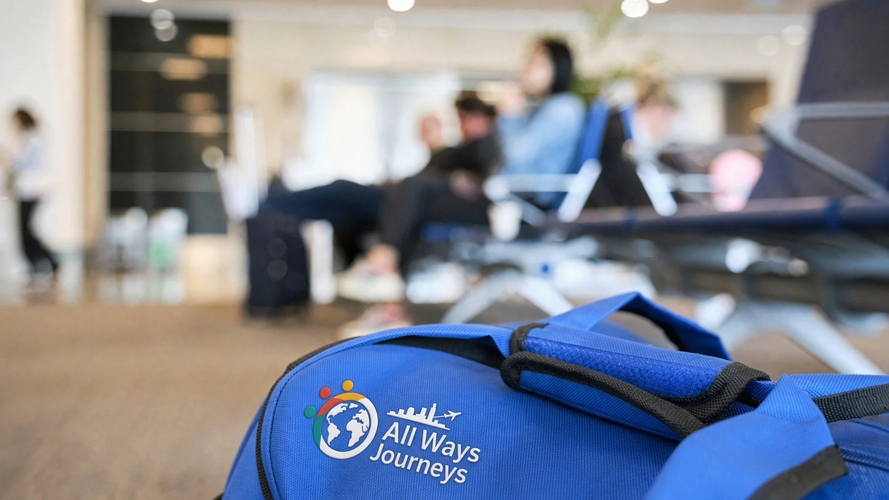 Close-up of a blue backpack with the logo and text 'All Ways Journeys' in an airport waiting area, with blurred passengers and seats in the background.