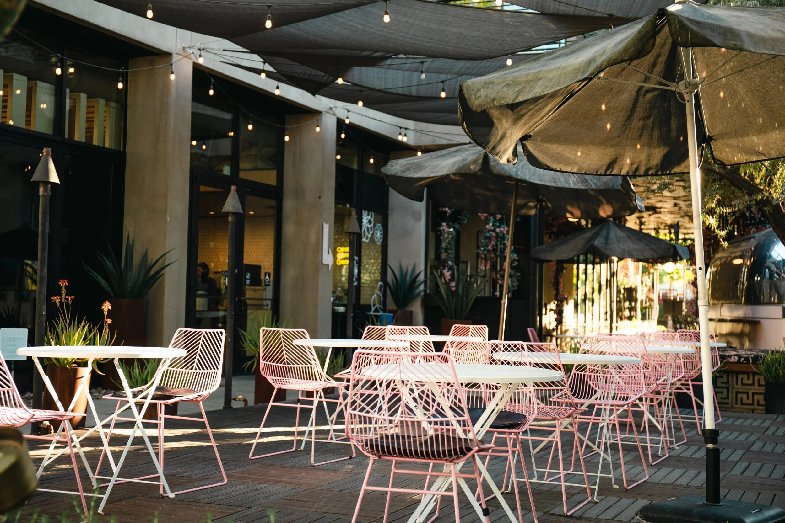 Outdoor patio seating area with pink wire chairs and white tables, umbrellas, string lights, plants, modern building with glass windows in the background.