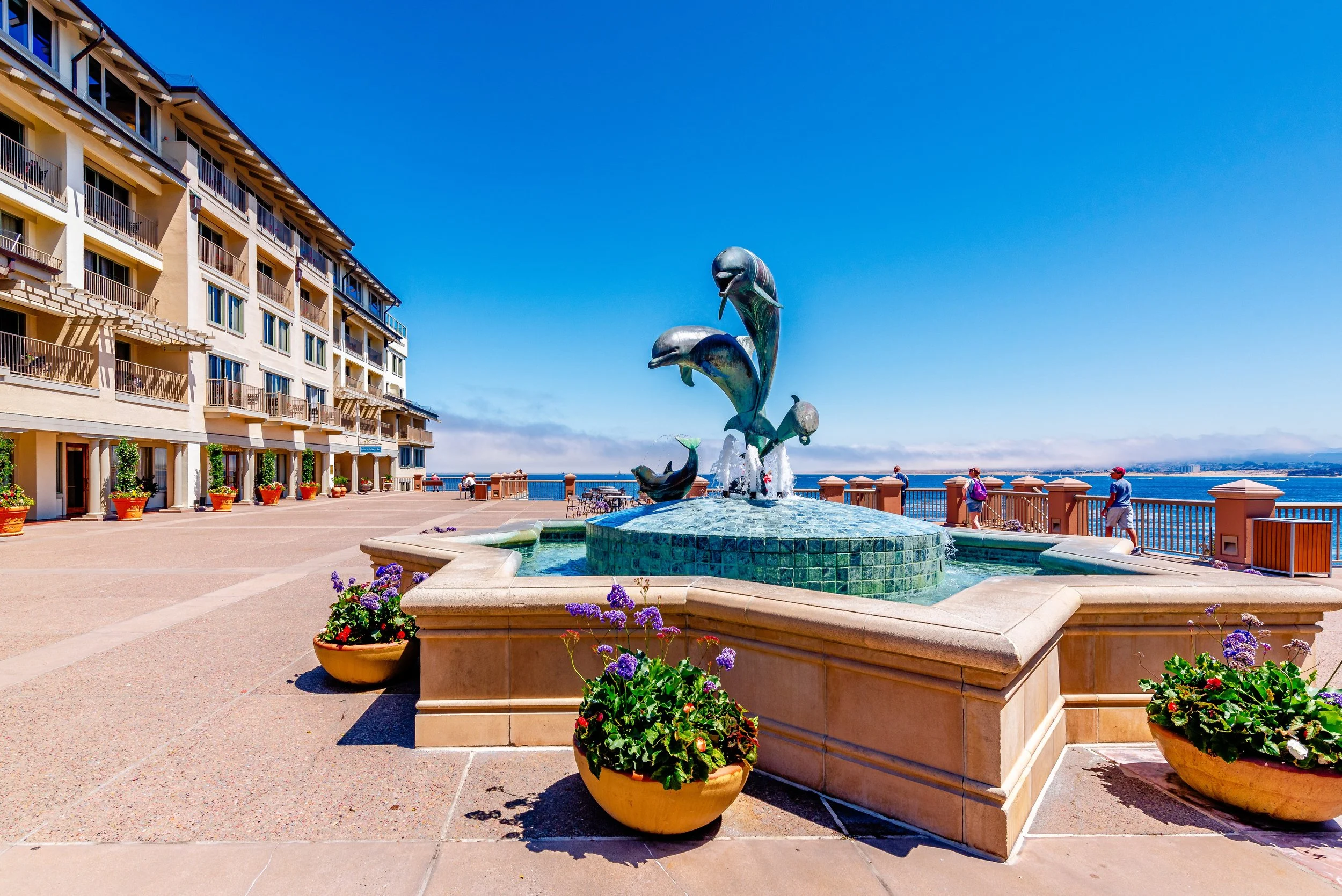 A seaside promenade with a fountain featuring dolphin sculptures, flower planters, a multi-story beige hotel with balconies on the left, and people walking along the railing overlooking the ocean under a clear blue sky.