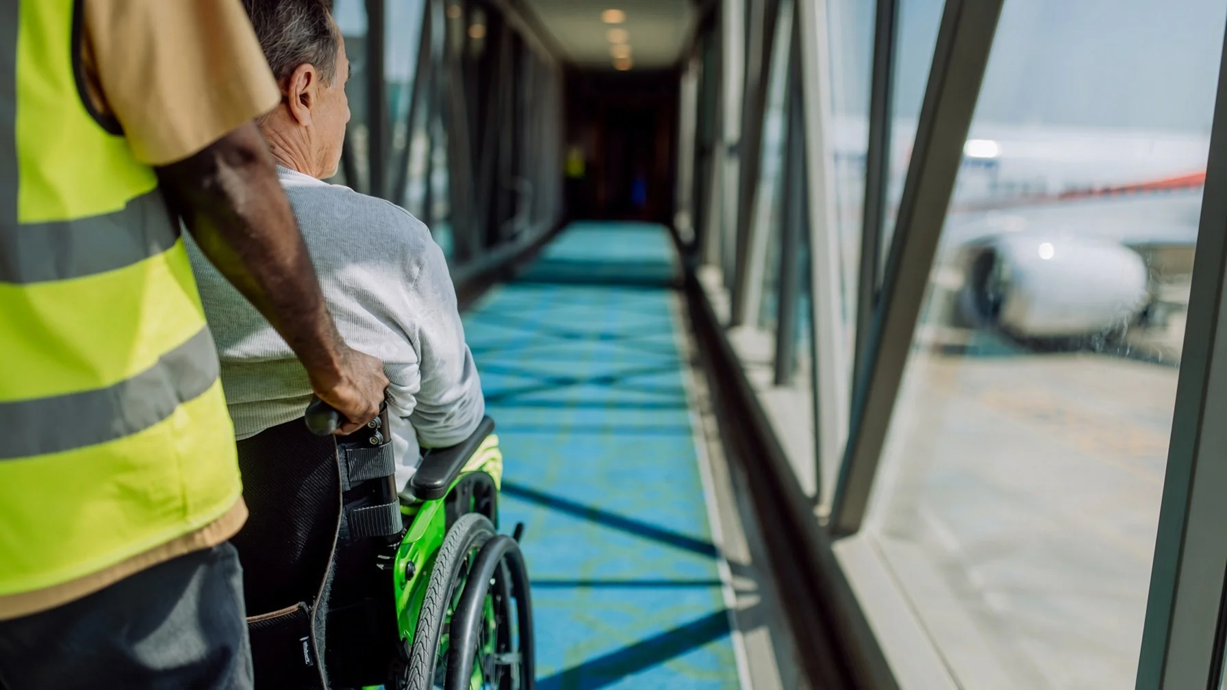 A person in a wheelchair being assisted by a staff member wearing a high-visibility vest at an airport gate, with airplane seen outside the large windows.