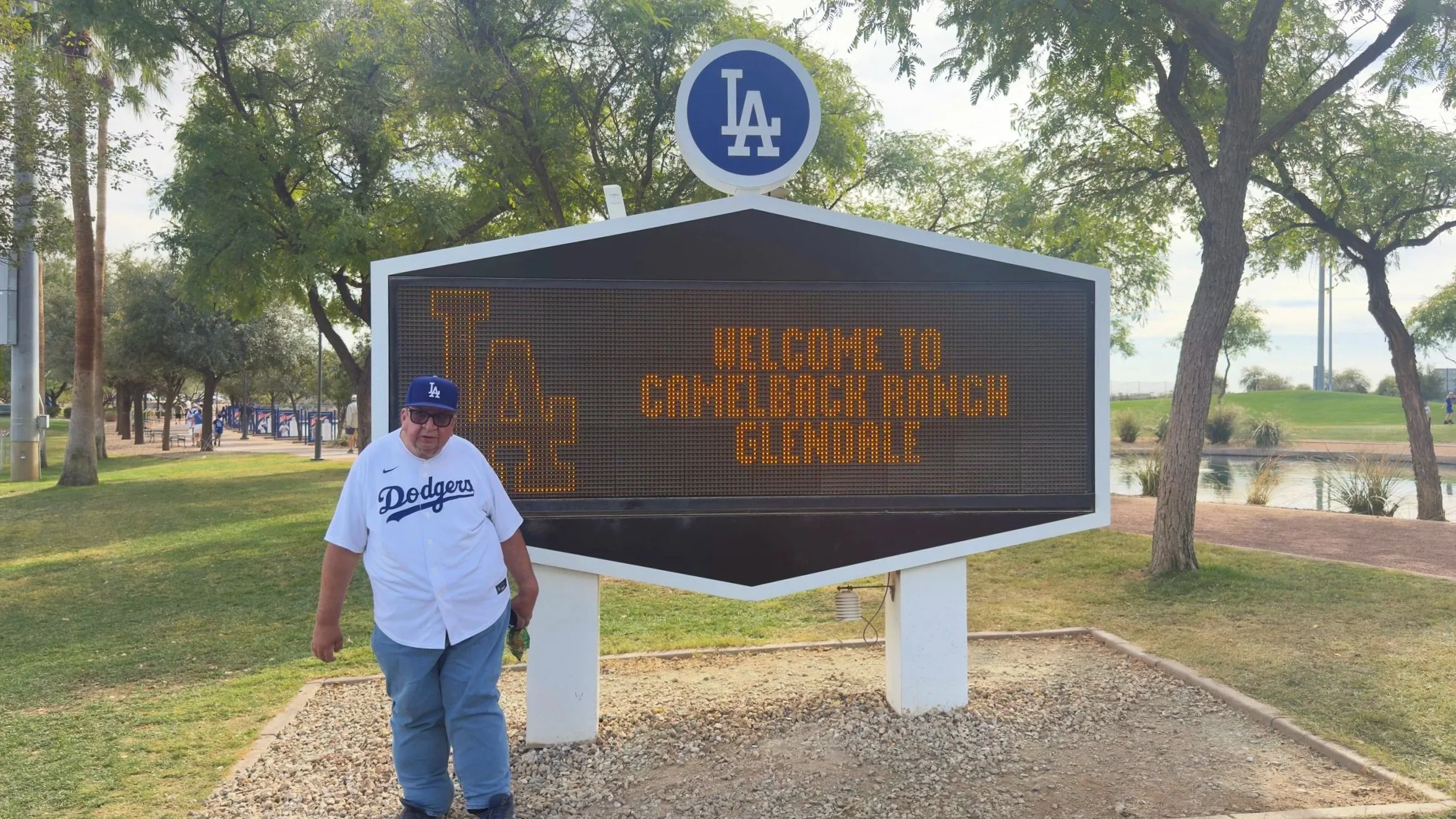 Man wearing a Dodgers jersey and cap standing in front of a digital sign that reads "Welcome to Camelback Ranch Glendale" at a park with trees and a pond in the background.