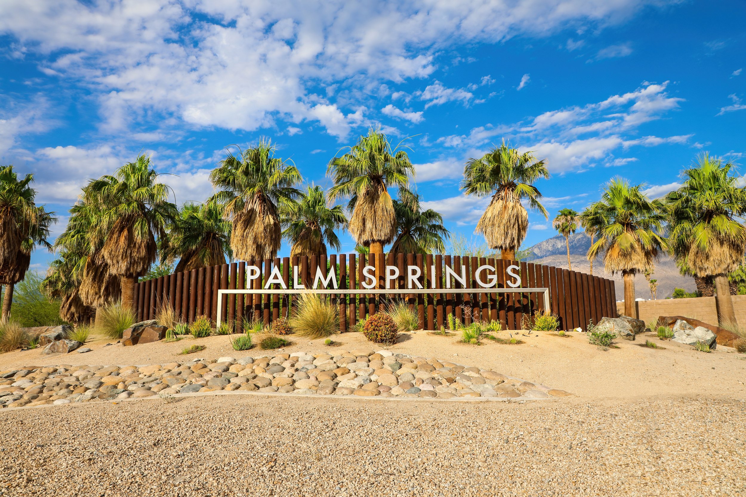 Sign reading 'Palm Springs' surrounded by palm trees, desert plants, rocks, and mountains in the background under a blue sky with scattered clouds.