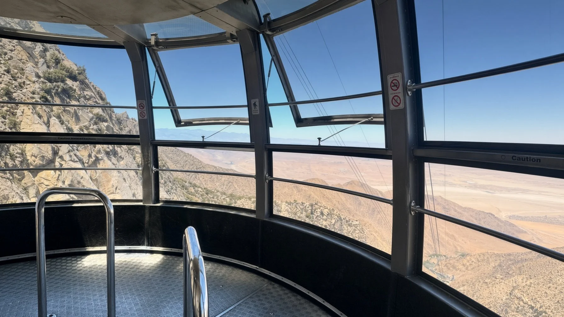 View from inside a glass-enclosed cable car or gondola with metal railings, showing rocky mountains and a valley landscape below, under a clear blue sky.