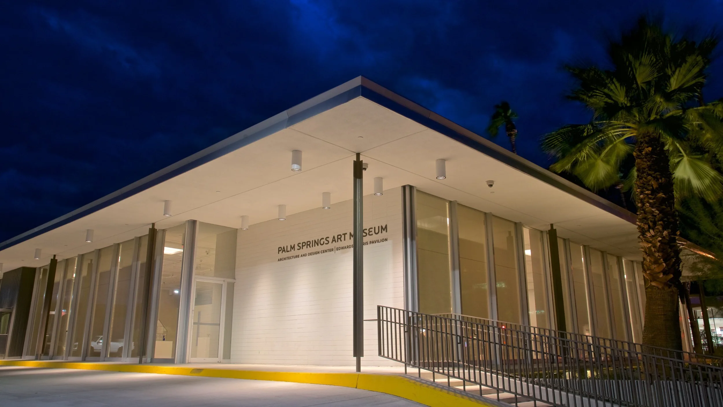 Night view of the Palm Springs Art Museum entrance with glass windows, palm trees, and a clear dark sky.