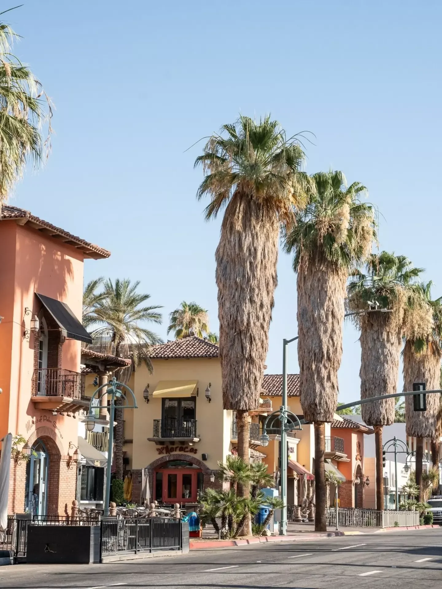A street scene with tall palm trees, colorful buildings with Spanish-style architecture, and outdoor seating areas in a warm, sunny climate.
