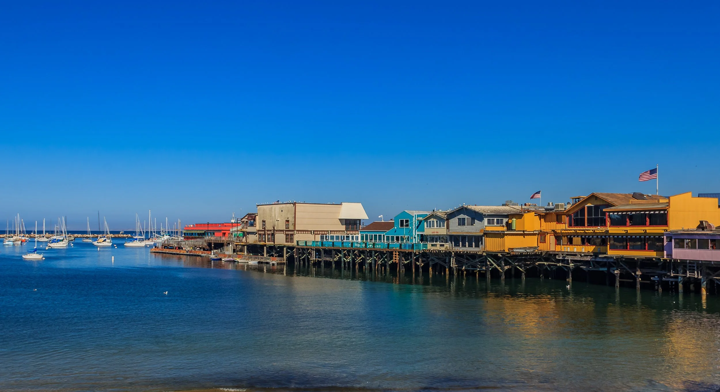 Colorful waterfront buildings and houses on a pier with boats anchored in the water and American flags flying, under a bright blue sky.