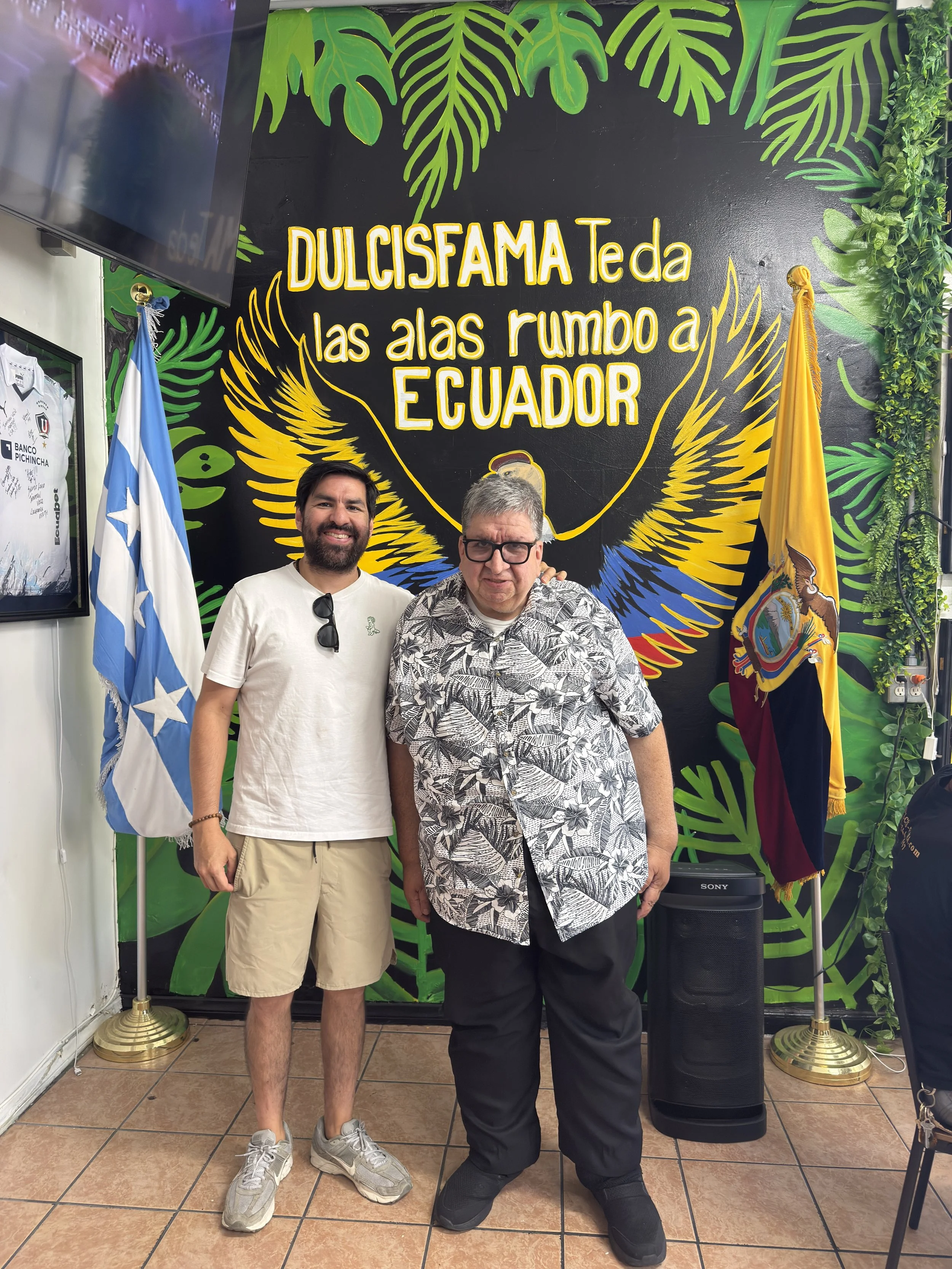 Two men standing in front of a colorful mural with large green leaves and yellow text that reads "DULCISFAMA Teda las alas rumbo a ECUADOR." They are smiling and standing on a tiled floor, with flags on either side and a black speaker nearby.