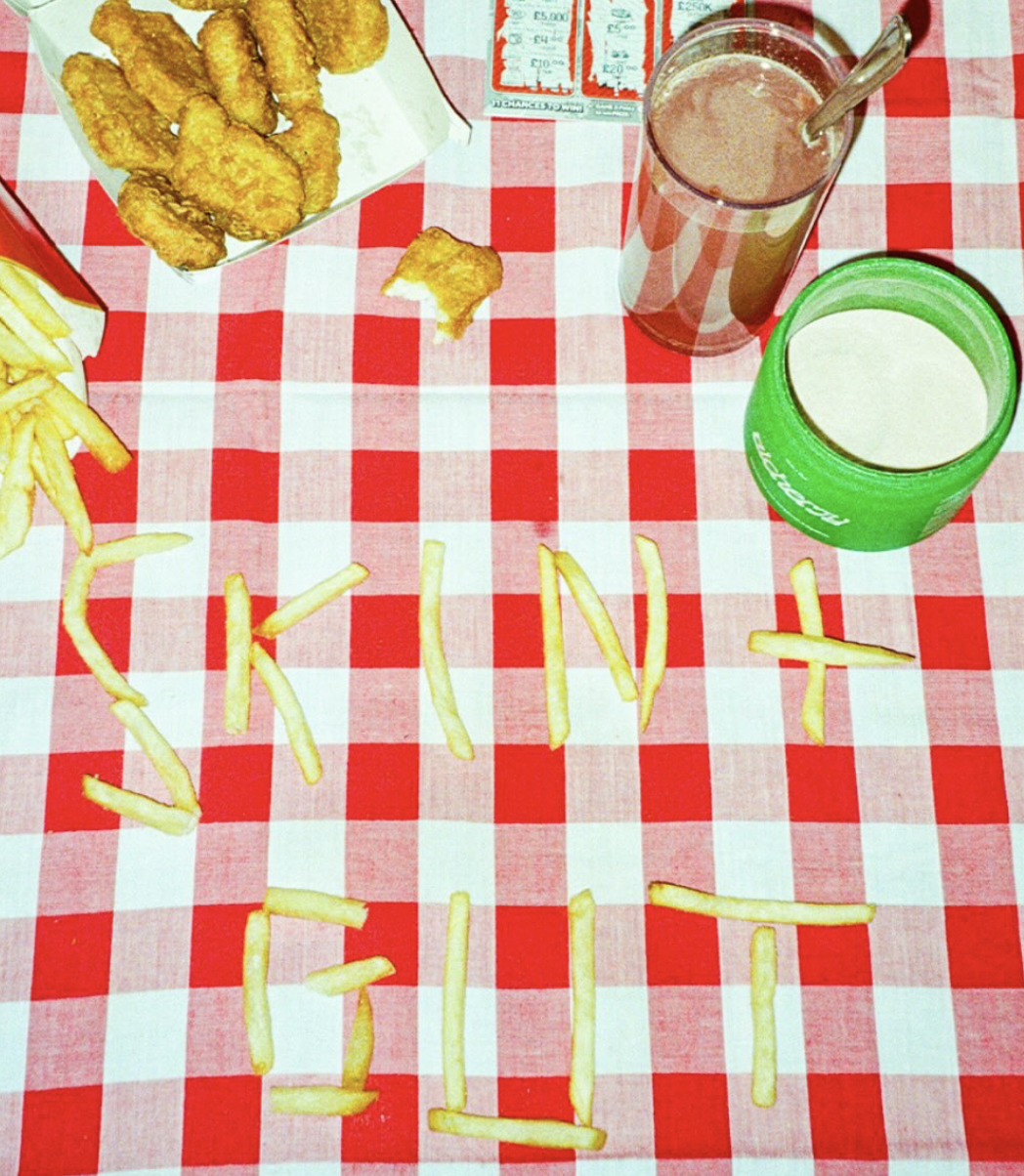 A checkered red and white tablecloth with scattered French fries, a paper food container with fried chicken pieces, two drinks in cups, and the words 'Thank You' spelled out with French fries.