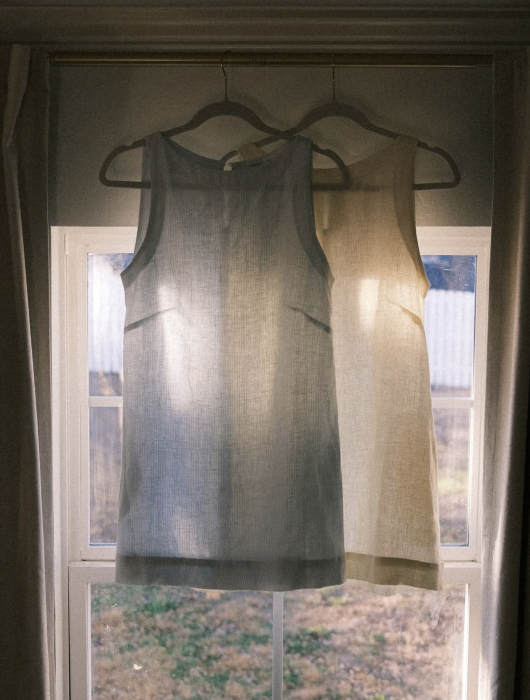 Two sleeveless linen dresses hanging on hangers inside a window frame.