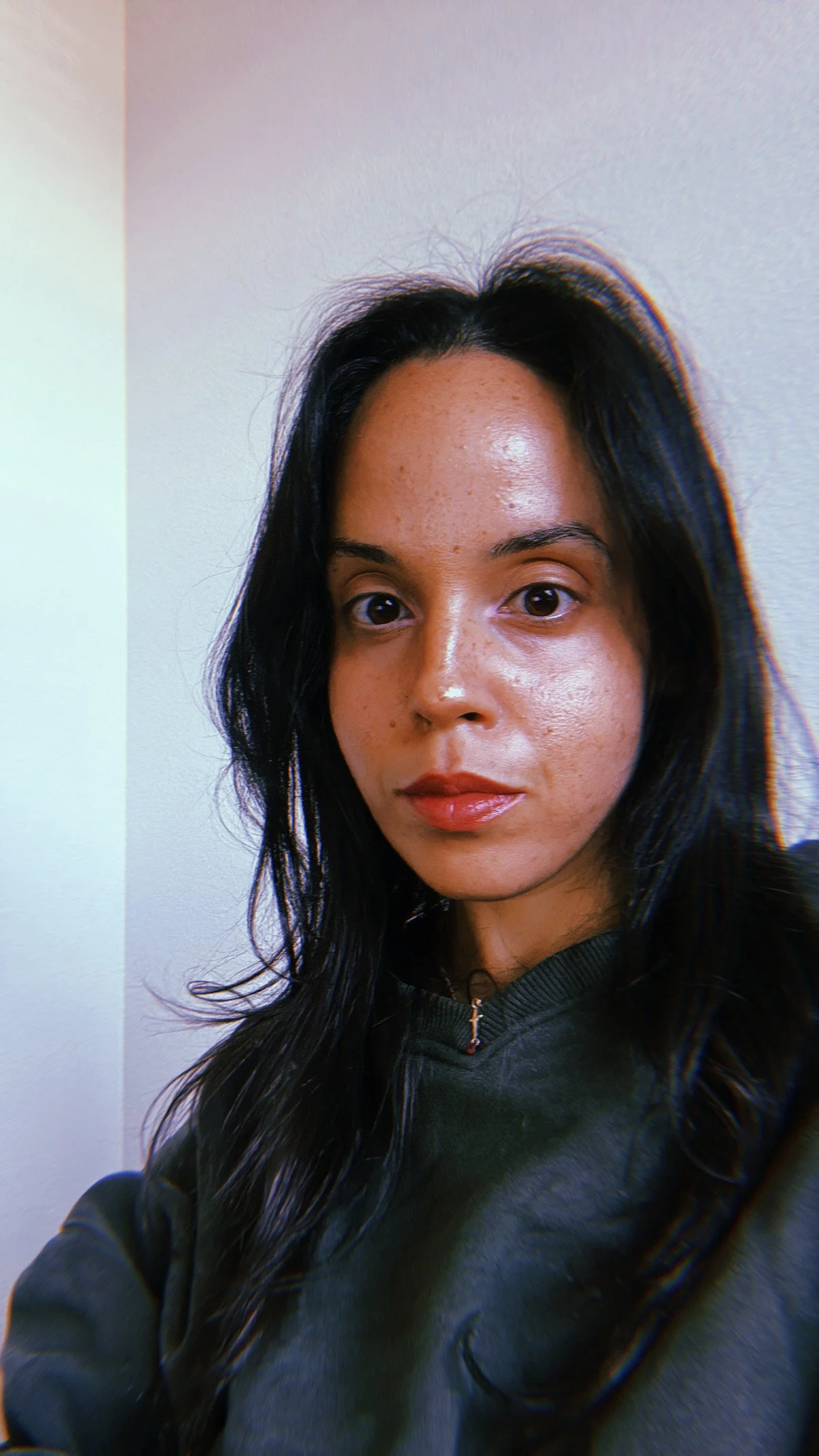 A woman with long black hair and a natural complexion taking a selfie indoors, wearing a dark jacket.