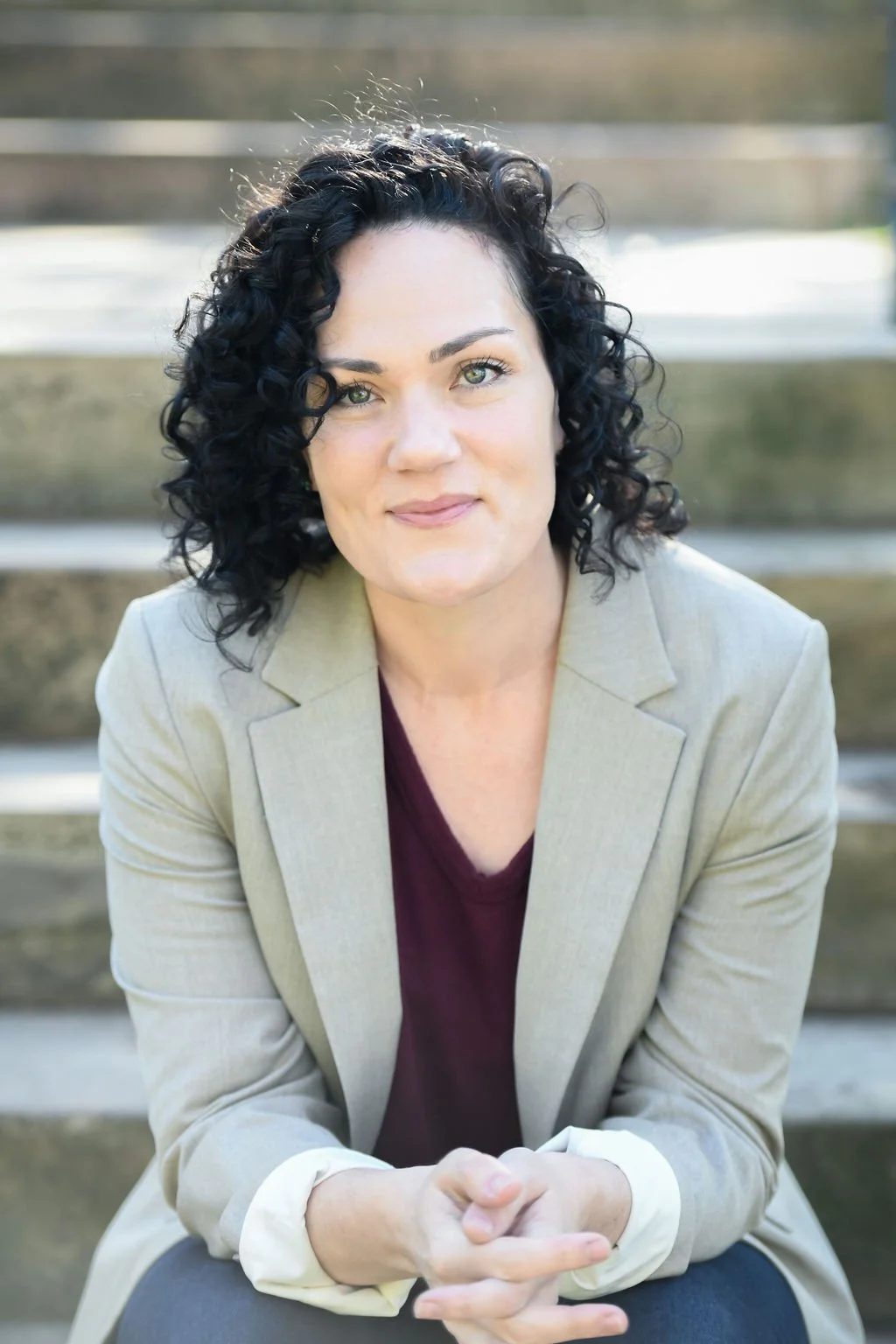 A woman with curly black hair, light skin, and green eyes, sitting on outdoor steps, wearing a beige blazer over a maroon top, looking at the camera.