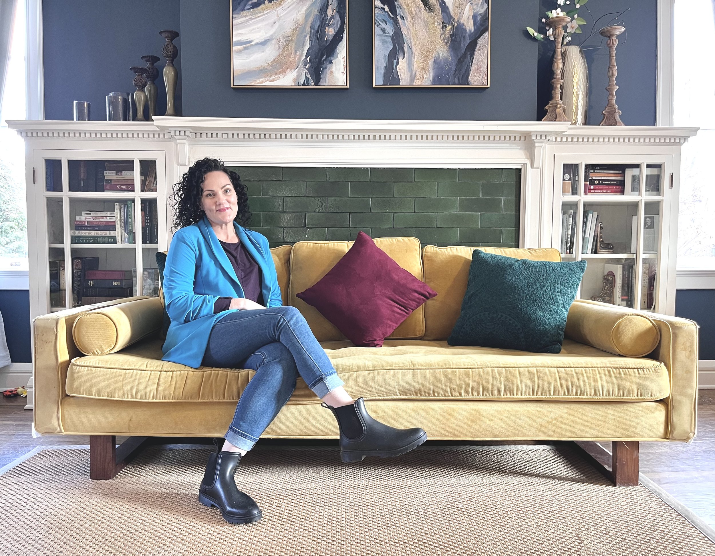 A woman with curly dark hair sitting on a yellow sofa in a living room. Behind her is a green brick fireplace with white built-in bookcases on either side. The sofa has a purple pillow and a teal pillow. The woman is wearing a bright blue blazer, a dark purple top, jeans, and black ankle boots.