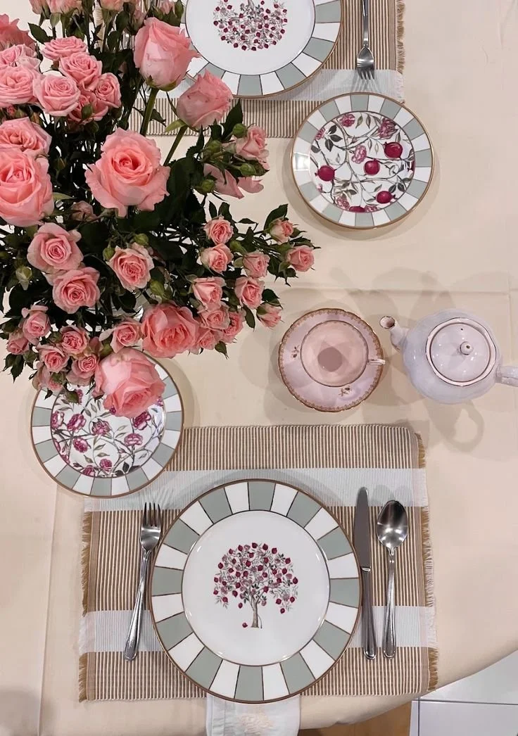 A dining table set with pink roses in a vase, decorative plates with floral and cherry designs, a teapot, a cup, and utensils on a striped placemat.