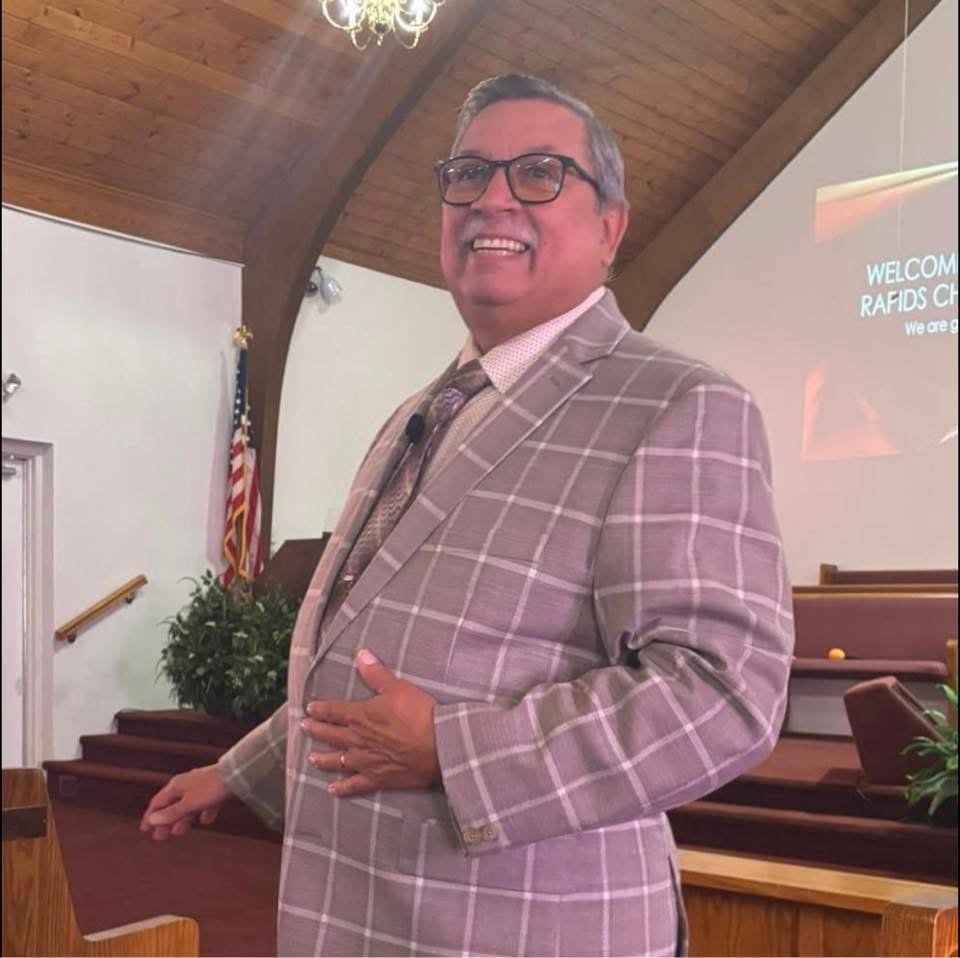 A middle-aged man with gray hair, glasses, and a broad smile, dressed in a beige plaid suit, standing inside a church or chapel.