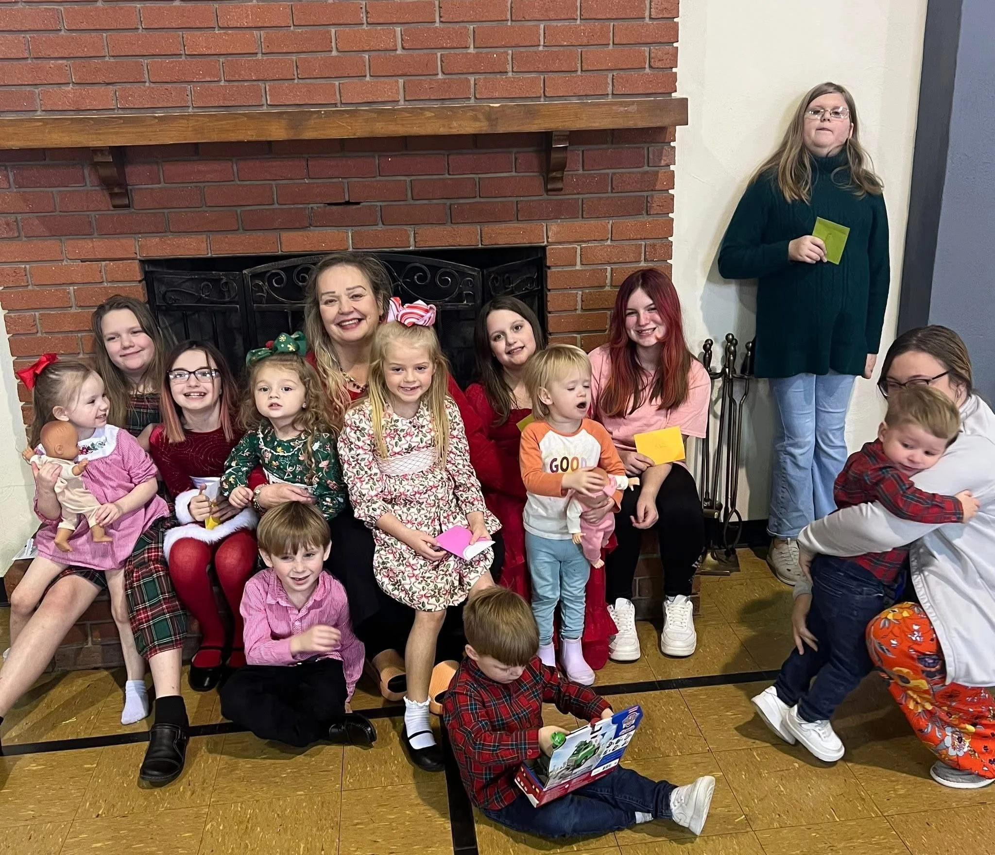 Group of children and adults, smiling, celebrating Christmas, sitting and standing in front of a brick fireplace, dressed in holiday clothes, some holding gifts or toys.