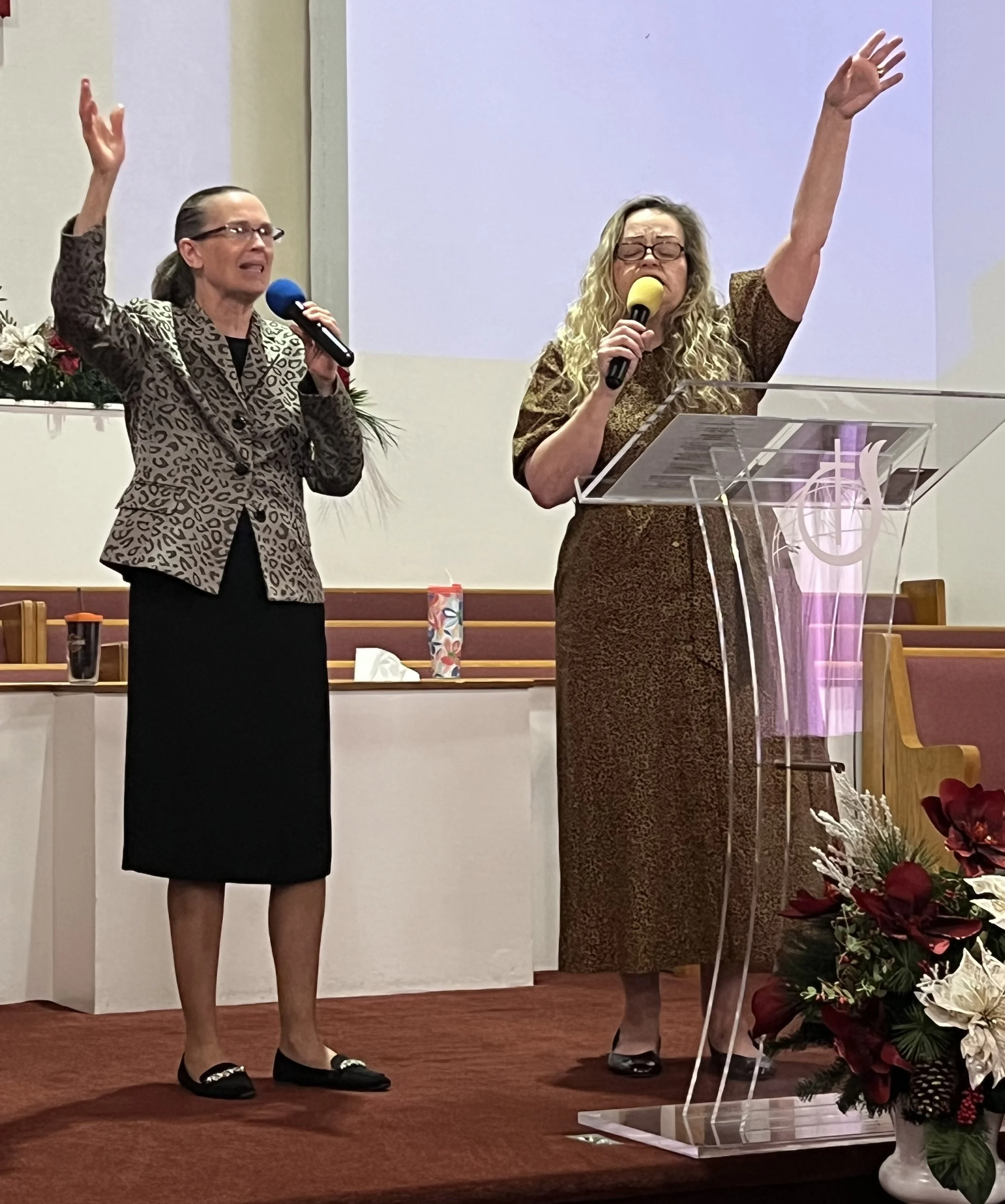 Two women standing on a stage, each holding a microphone and appearing to sing or speak passionately with their eyes closed. One woman has dark curly hair and wears a leopard print blazer with a black skirt, while the other has curly blonde hair and wears a brown patterned dress. There is a clear lectern in front of the blonde woman, and floral arrangements are visible in the foreground.