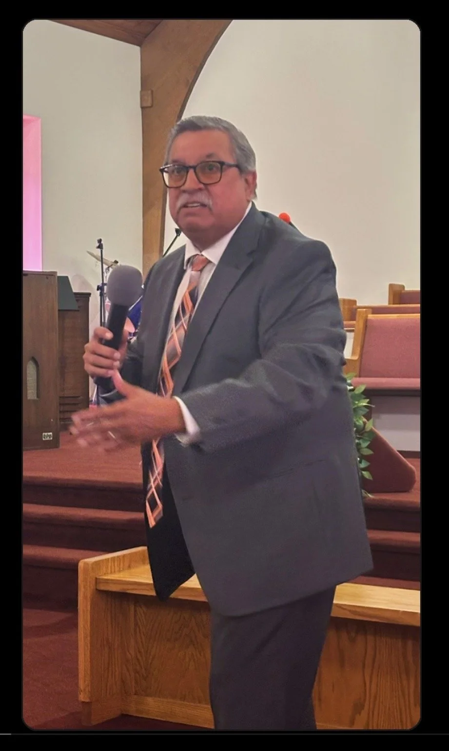 A man in a gray suit, white shirt, and colorful tie holding a microphone, speaking inside a church or auditorium with wooden pews and a curved white wall in the background.