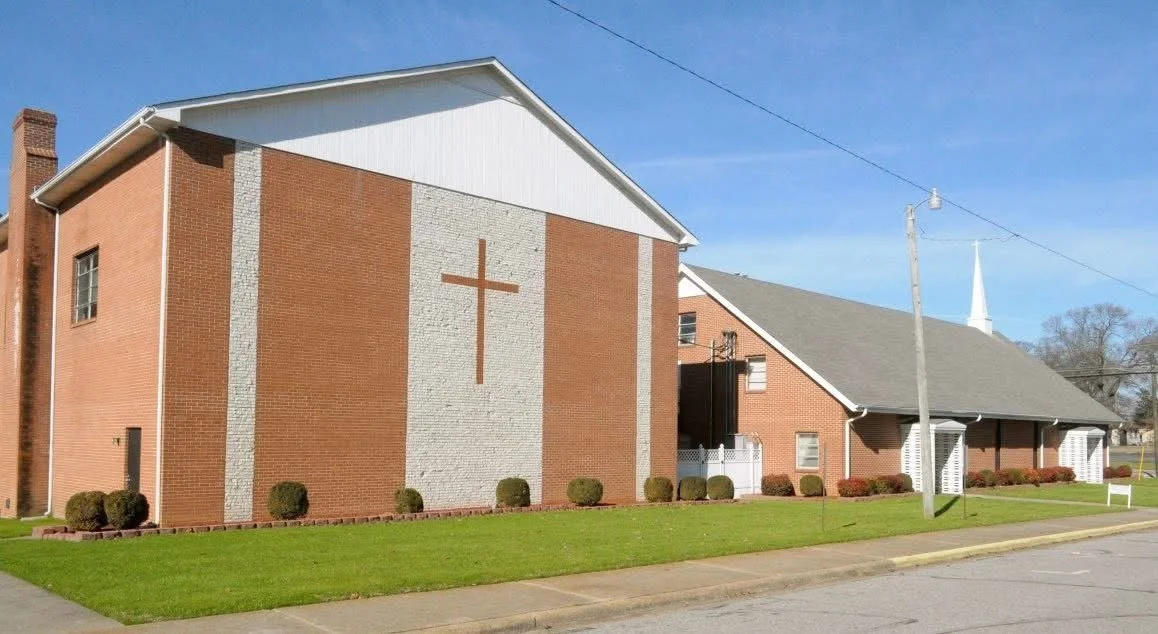 A brick church with a large cross on the front facade, a grassy lawn, and a sidewalk in front, under a clear blue sky.