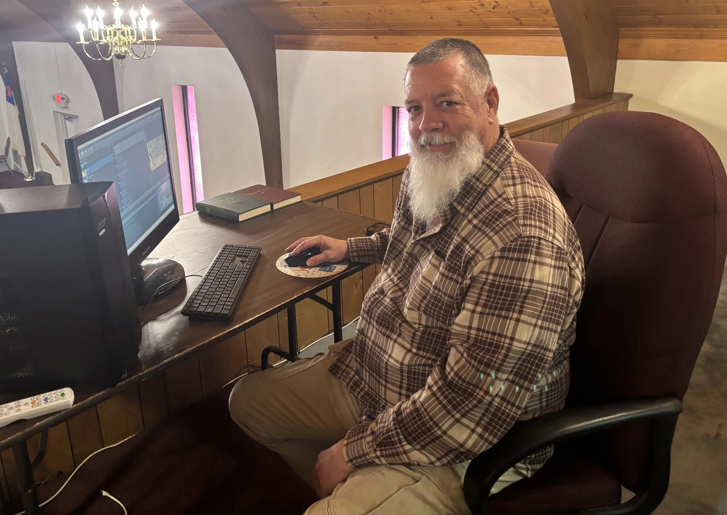 A man with a white beard and gray hair, wearing a plaid shirt, sitting at a desk with a computer in a room with wooden accents, chandelier, and pink vertical light fixtures.