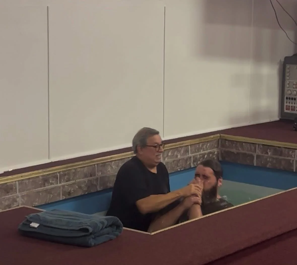 Two men are baptizing each other in a small baptismal pool in a church or religious setting. One man is holding the other man's head and nose under the water, while the second man is preparing for the baptism, with his hands clasped and eyes closed.