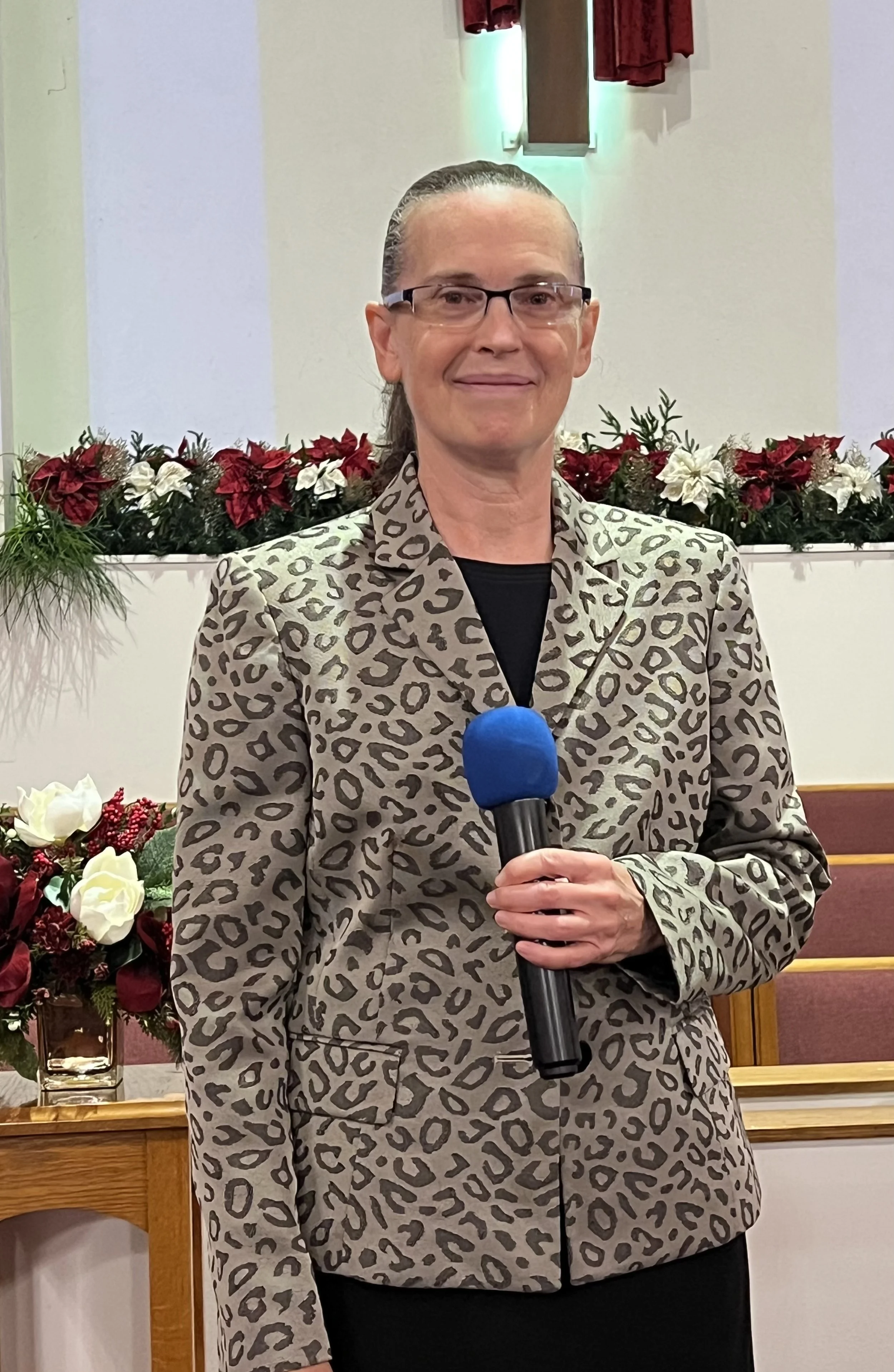 A woman wearing glasses and a leopard print blazer holding a blue microphone standing in front of Christmas decorations with poinsettias and greenery in a church or similar setting.