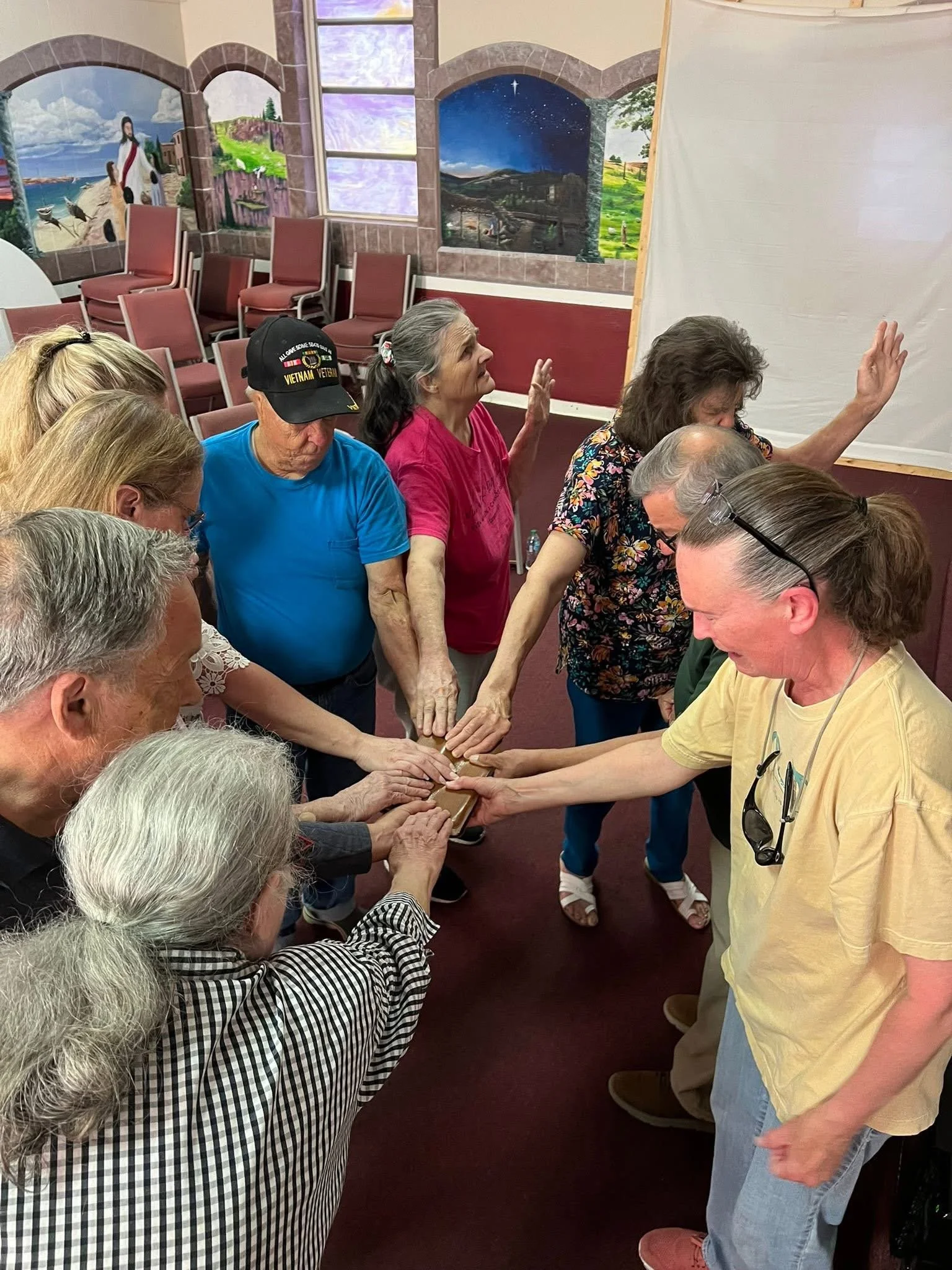 A group of elderly people and a caregiver participating in a group activity, with hands joined together in the center, inside a room with colorful mural paintings on the walls.