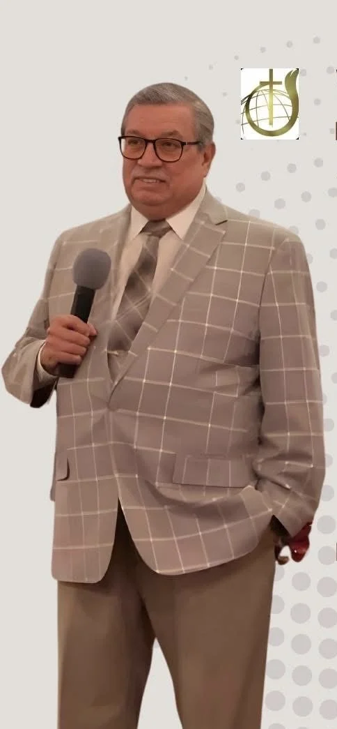 A man in a beige plaid suit holding a microphone and standing in front of a backdrop with a logo featuring a ukulele and musical notes.