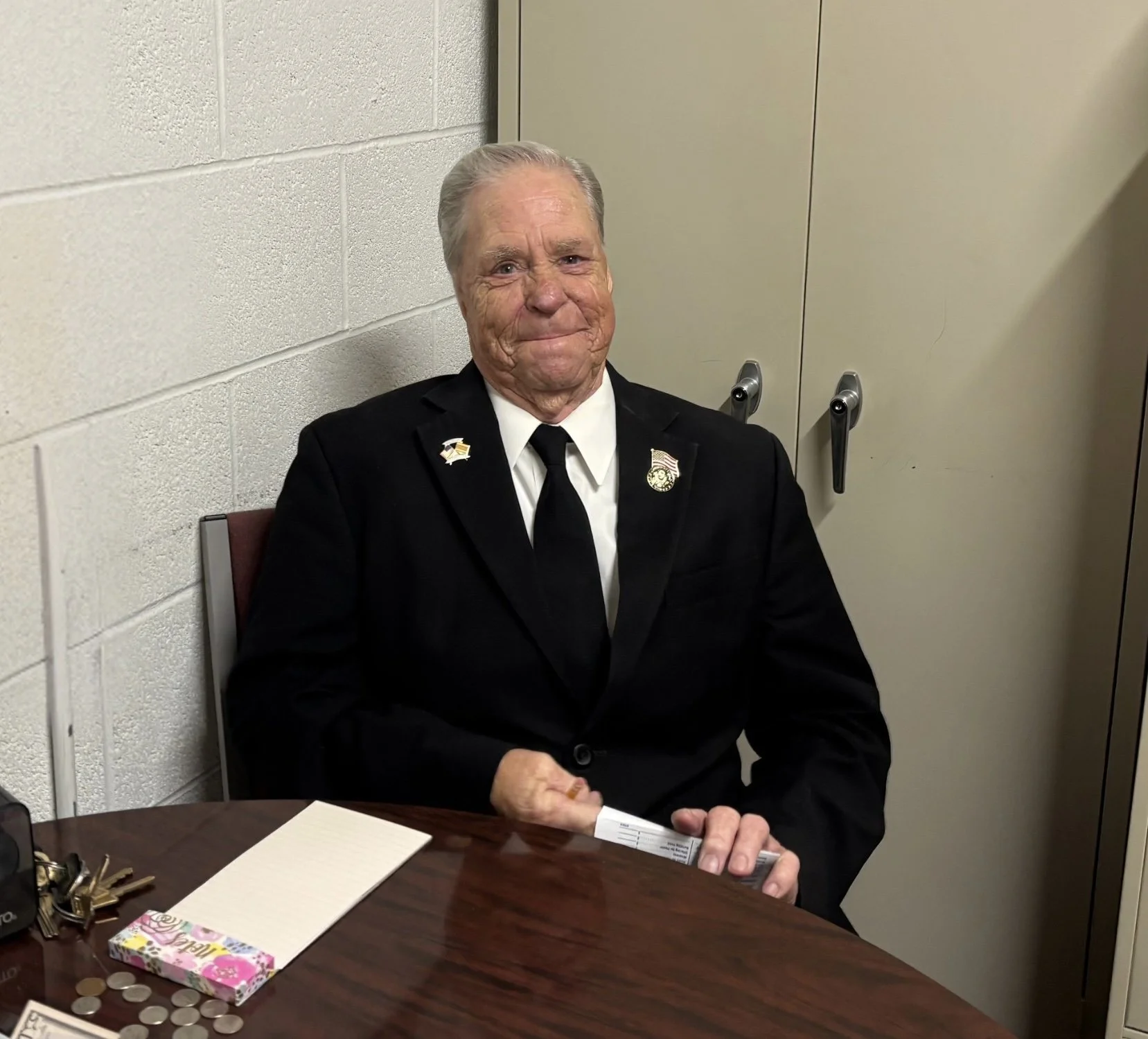 An elderly man dressed in a black suit with pins on his lapel, sitting at a table with a notepad, coins, and keys, in a room with beige walls and cabinets behind him.