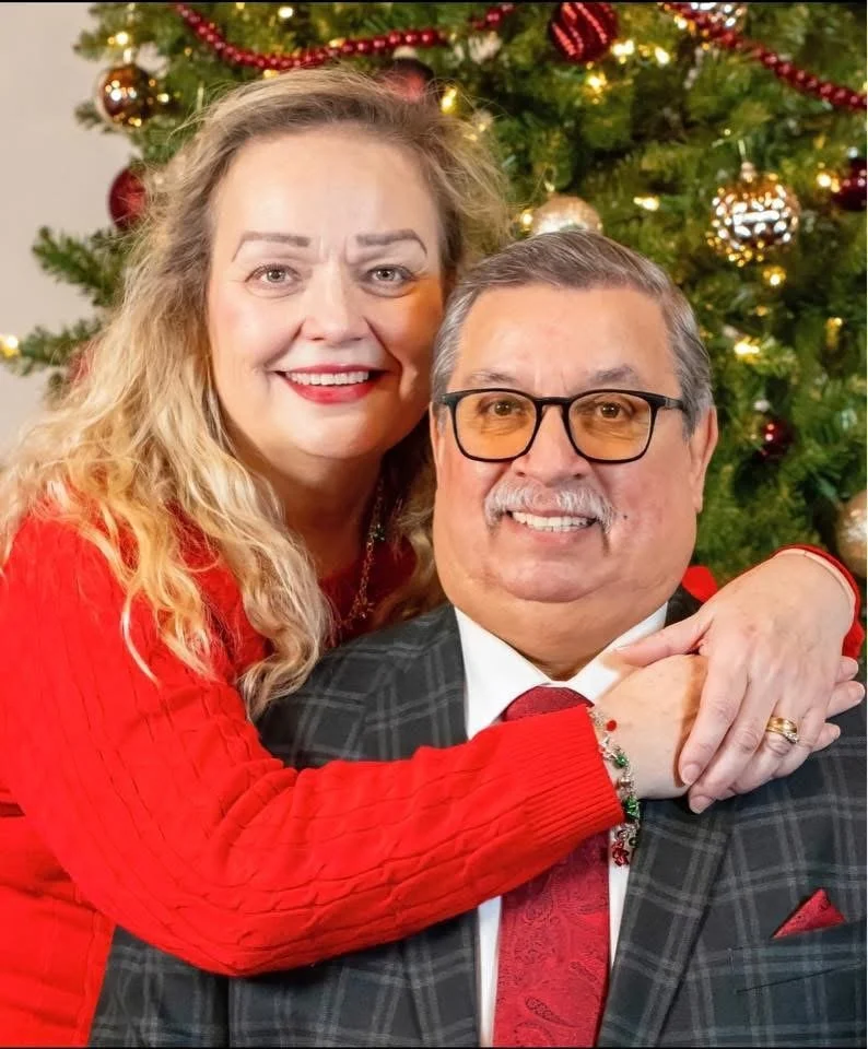 A smiling woman with curly blonde hair and a red sweater hugs a smiling man with glasses, a gray mustache, and a plaid suit in front of a decorated Christmas tree.