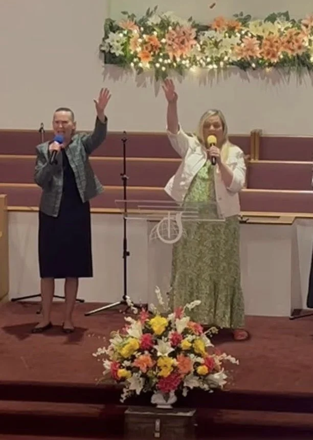 Two women are standing on a church stage with flowers, holding microphones, waving, and smiling at the audience.