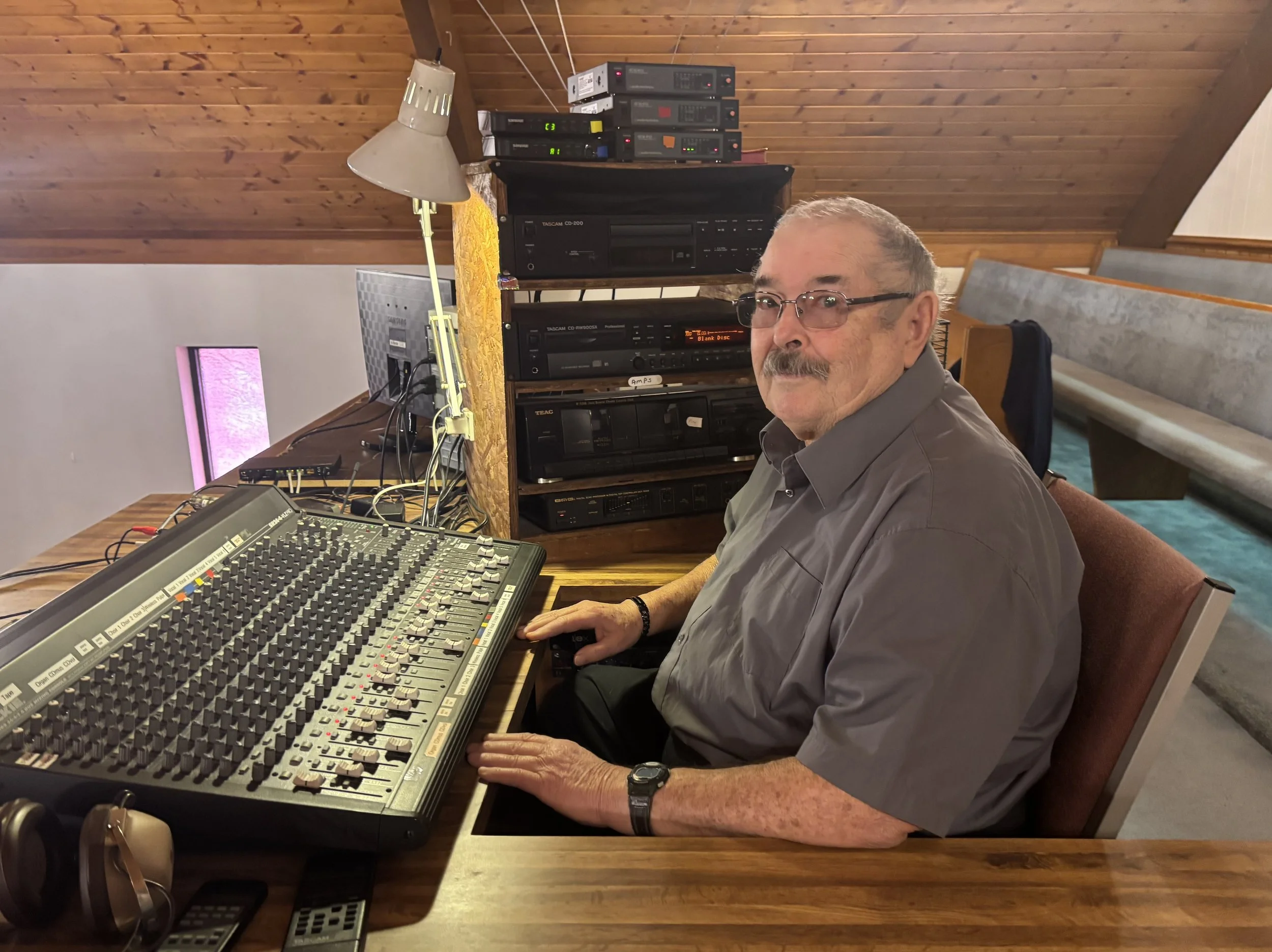 A man with glasses and a mustache, wearing a gray shirt, sitting at a mixing console in a room with wooden ceiling and walls, surrounded by audio equipment.