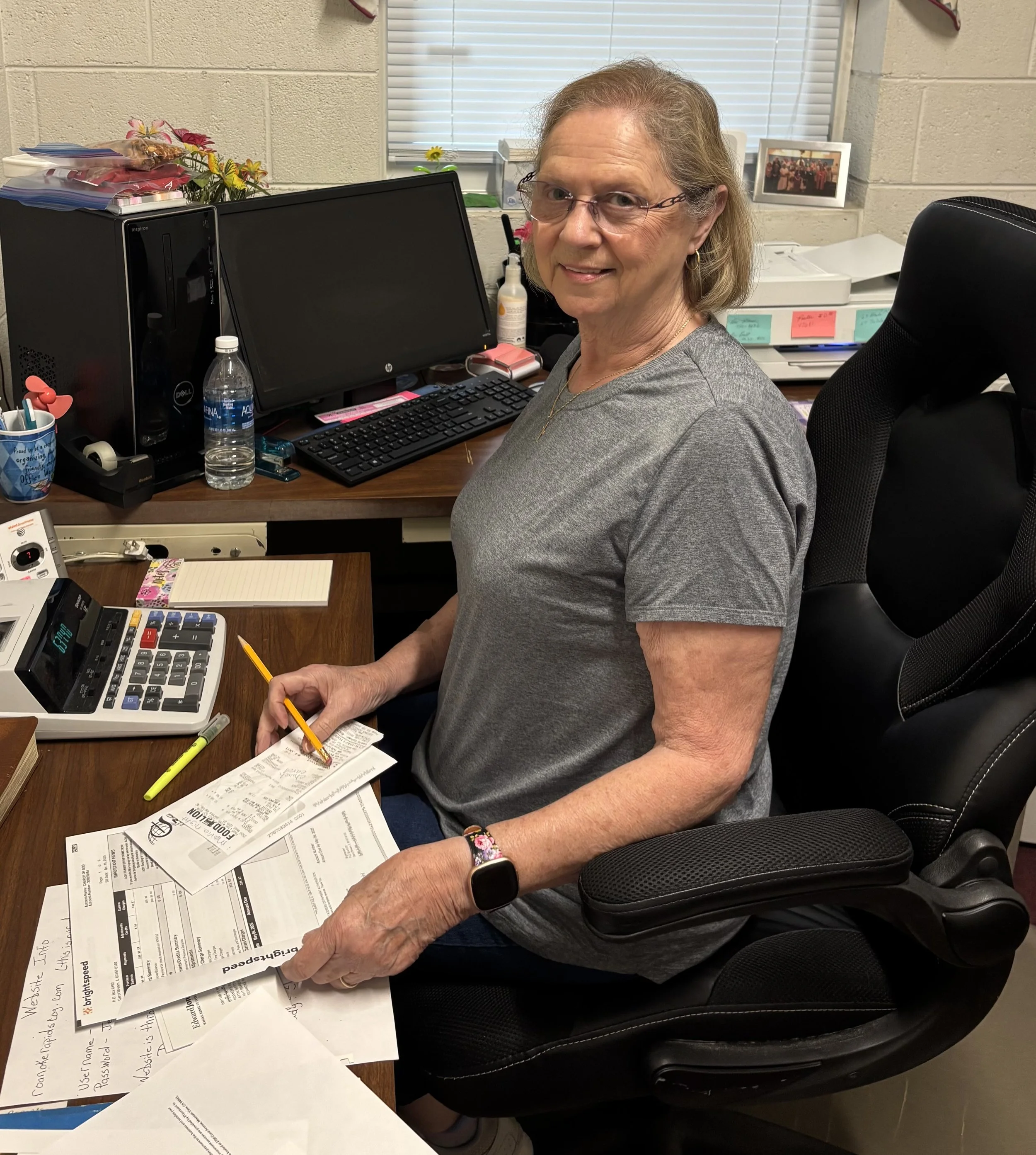 A woman sitting at a cluttered office desk, holding a receipt and writing on it, with papers, a calculator, computer monitor, and office supplies around her.