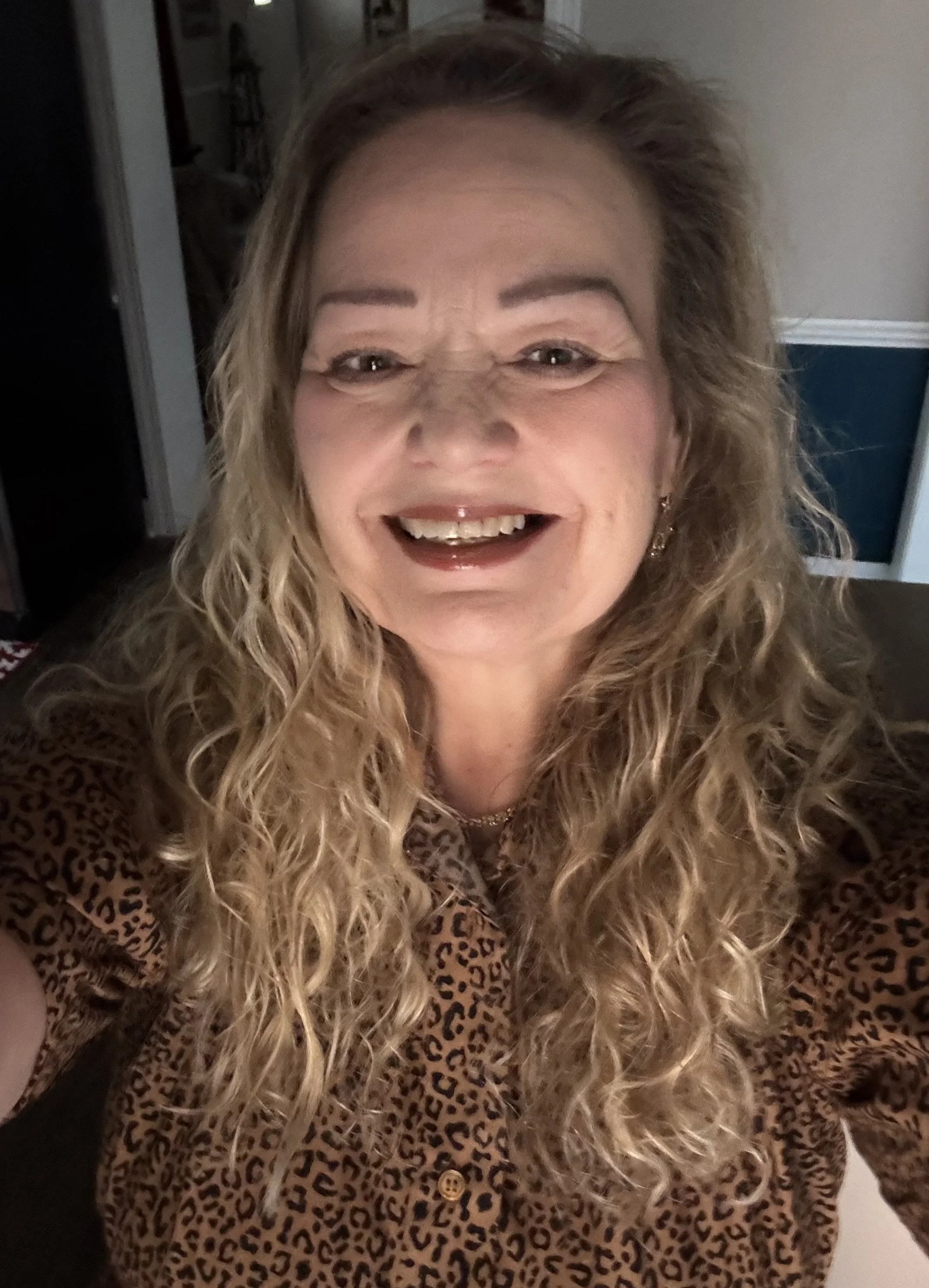 A woman with curly blonde hair taking a selfie, smiling, wearing a leopard print top, earrings, and a necklace, indoors with a room in the background.