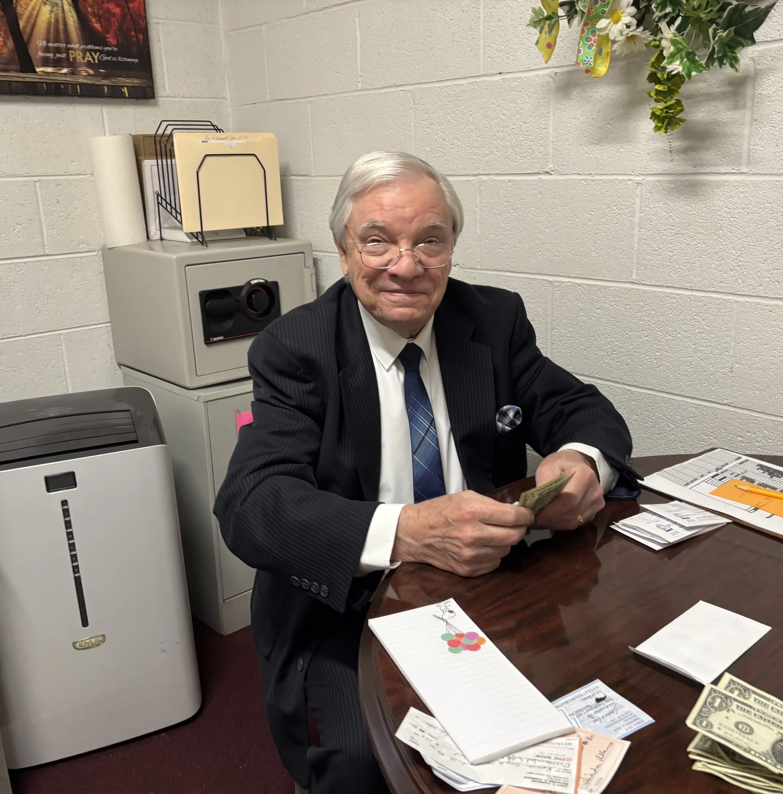 An elderly man with glasses, dressed in a black suit and tie, sitting at a desk, smiling, and holding cash in his hand, in an office setting with papers, a notepad, and a package of snacks on the desk.