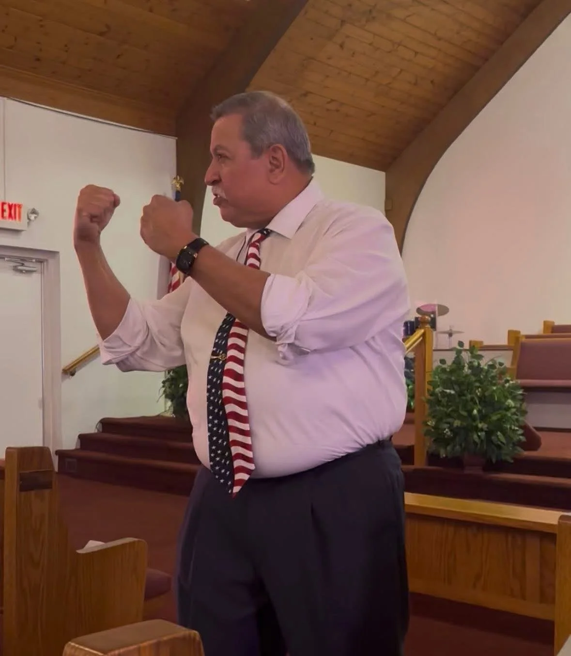 A middle-aged man in a white dress shirt with rolled-up sleeves, a patriotic American tie, and dark pants stands inside a church. He is clenching his fists and has an intense expression, possibly speaking passionately. The church interior features wooden pews, an altar with plants, and an American flag in the background.