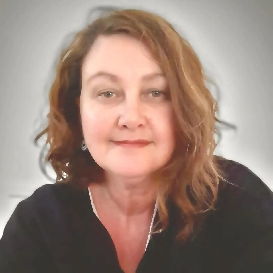 A woman with curly red hair, wearing earrings and a black top, looking at the camera against a plain background.