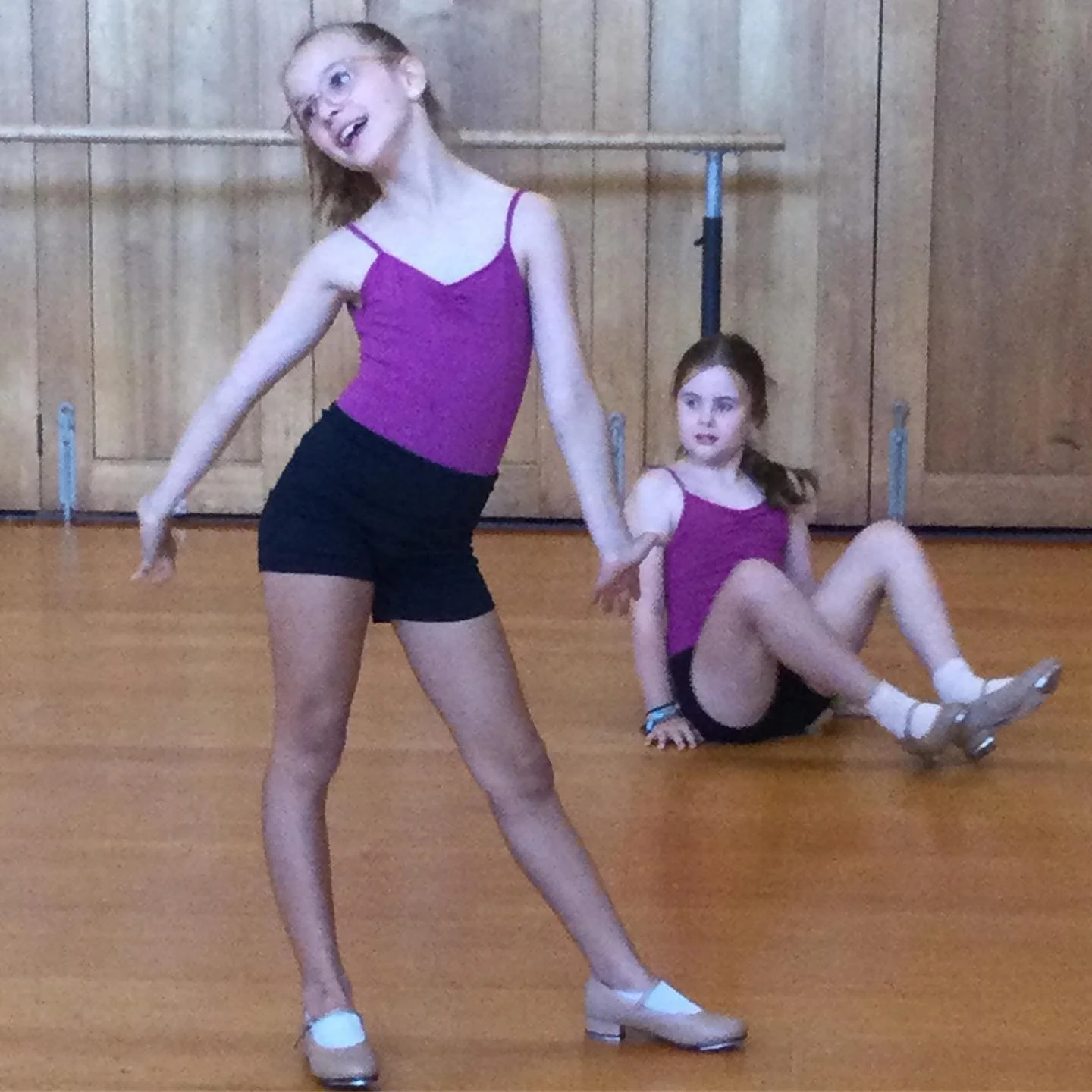 Two young girls in dance costumes practicing in a dance studio with wooden walls and flooring. One girl is standing in a pose, the other girl is sitting on the floor.