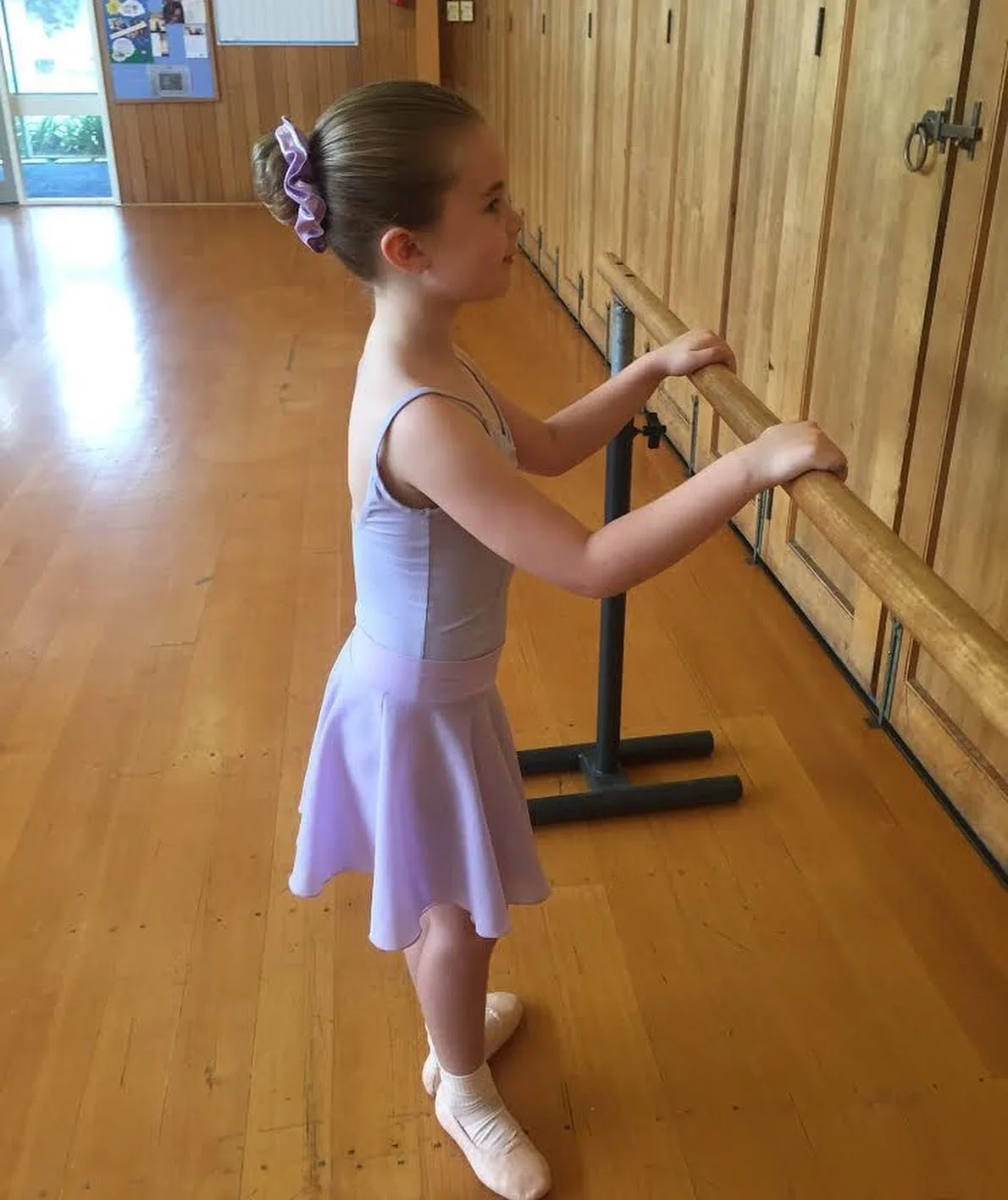 Young girl in a pink ballet dress and ballet shoes standing at a ballet barre in a dance studio.