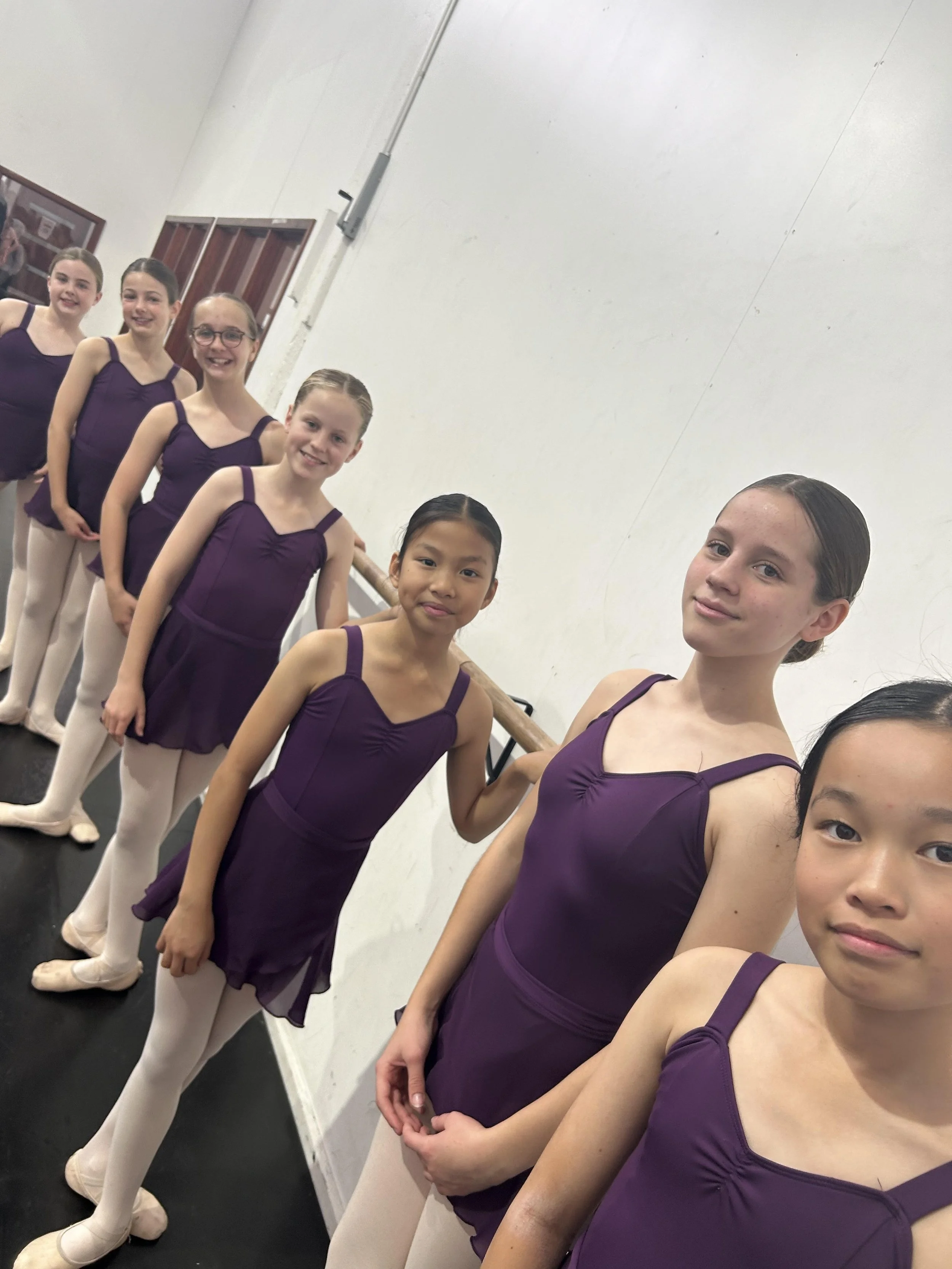 A line of young female ballet dancers in purple leotards and white tights, standing at the barre inside a dance studio.
