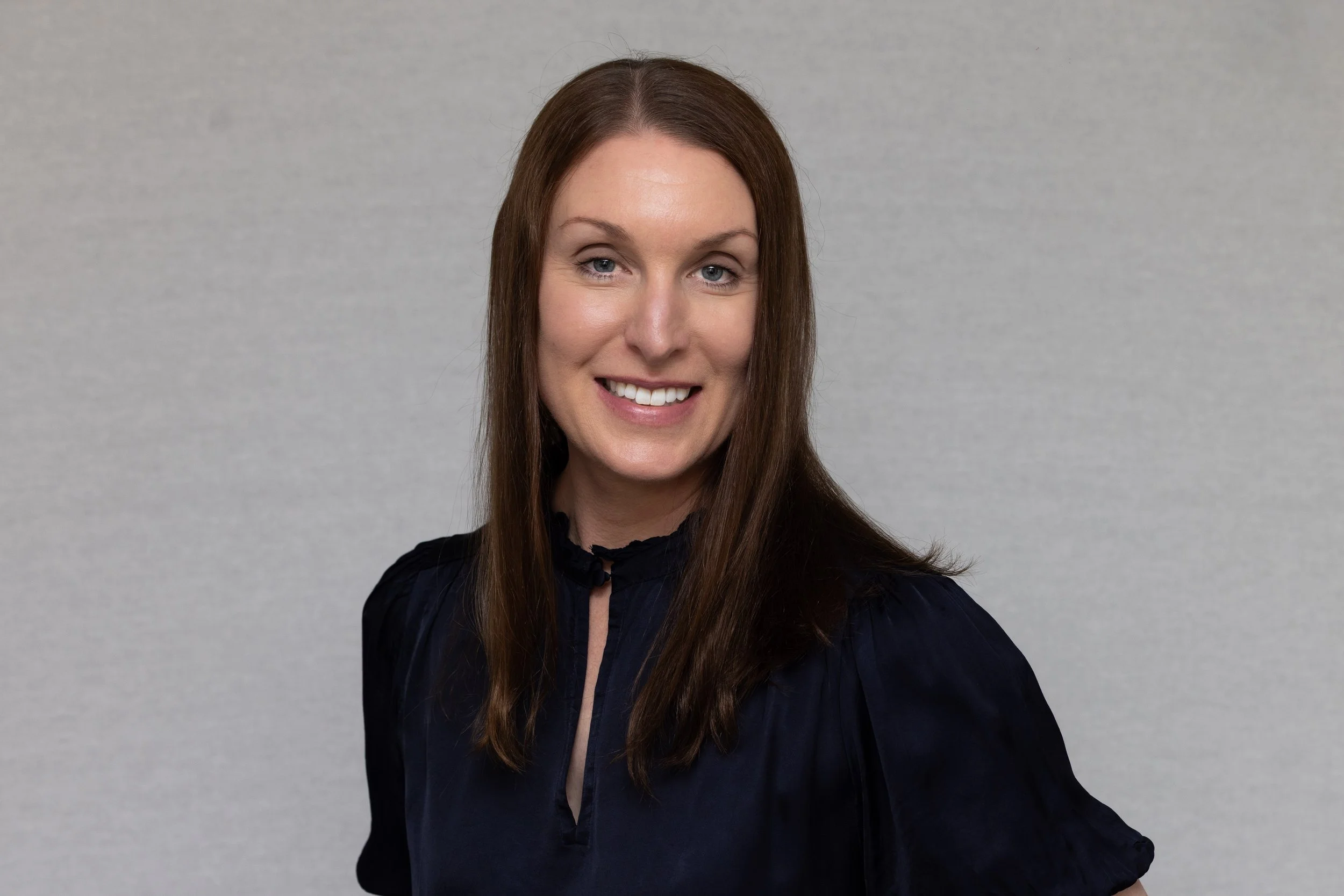 Portrait of a woman with light skin, long brown hair, and blue eyes, smiling against a plain light gray background.