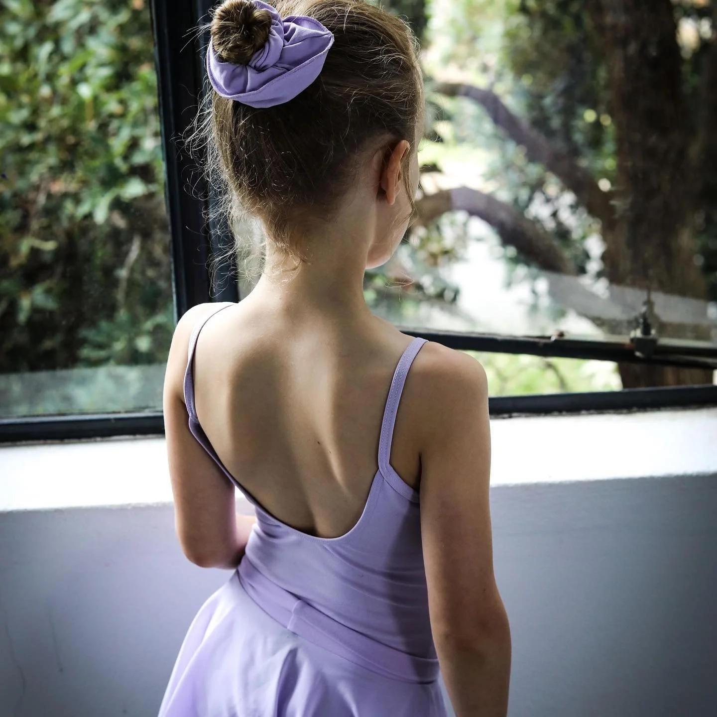 A young girl with brown hair tied up with a purple scrunchie, wearing a lavender dress, looking out a window at a large tree outside.