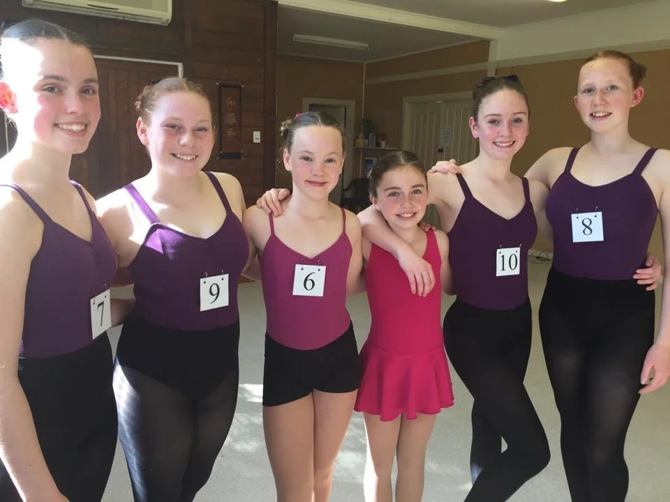 Six young girls in ballet costumes standing together in a room, wearing numbered tags with numbers 7, 9, 6, 5, 10, and 8, smiling at the camera.